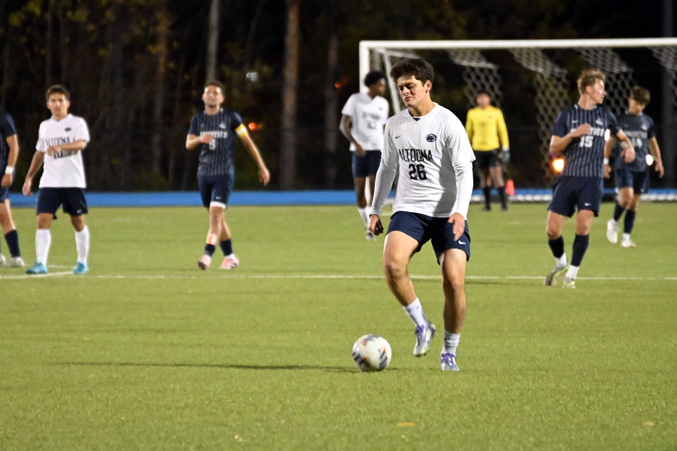 A young male soccer player wearing a white jersey with the number 26 and the word 'Penn State' on it, is walking on a soccer field at night with a soccer ball at his feet. There are other players in the background, some in white and others in navy bl
