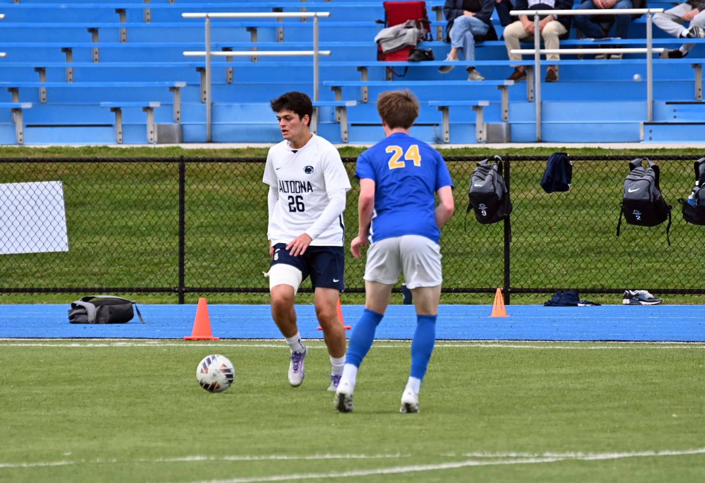 Two young soccer players, one in a white jersey and the other in a blue jersey with the number 24, are on the field during practice or a game. Blue bleachers and a few seated spectators are visible in the background.