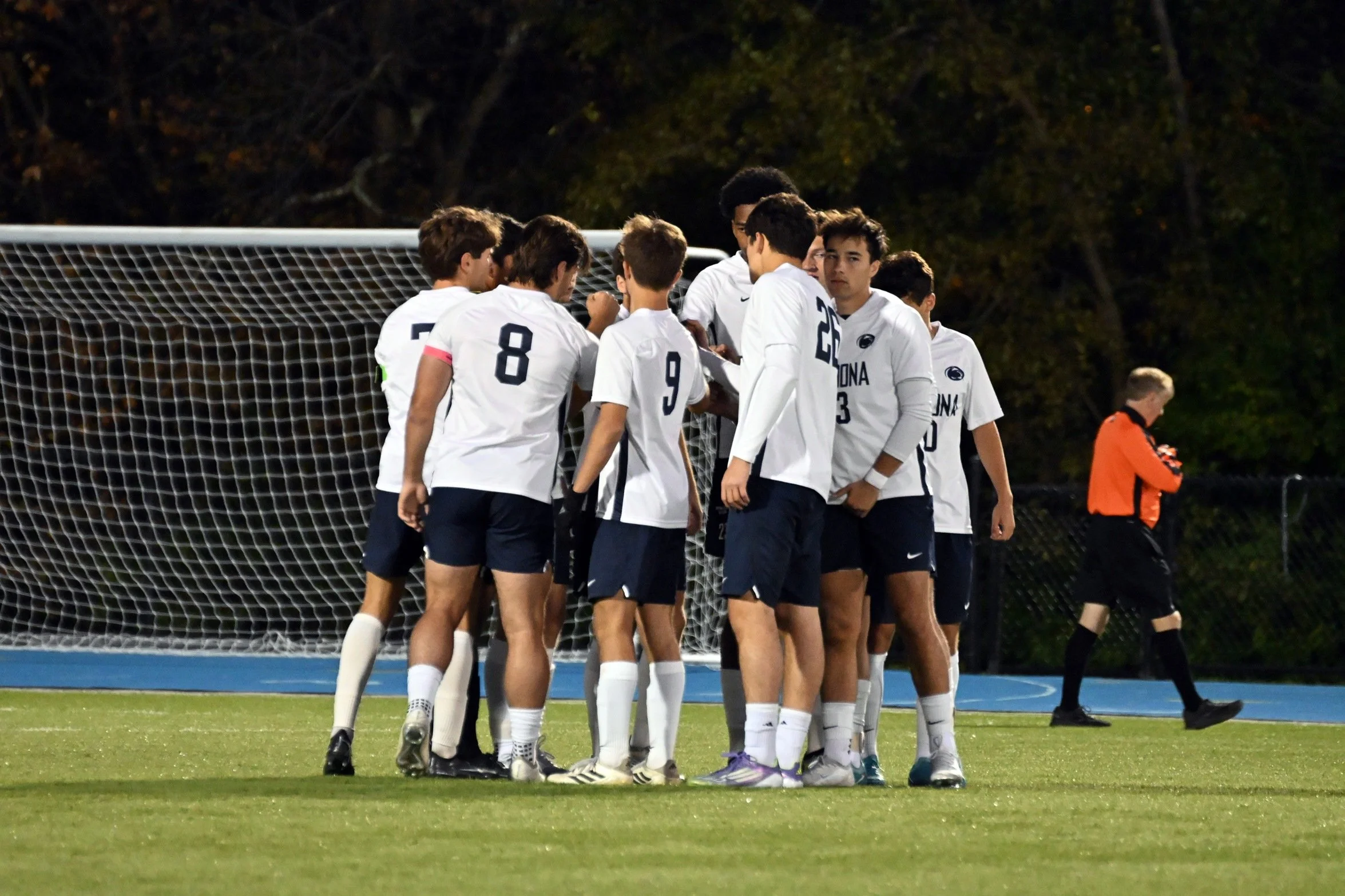 A group of young male soccer players in white jerseys gathers around near the goalpost on a soccer field at night, with an official or coach in an orange jersey in the background.