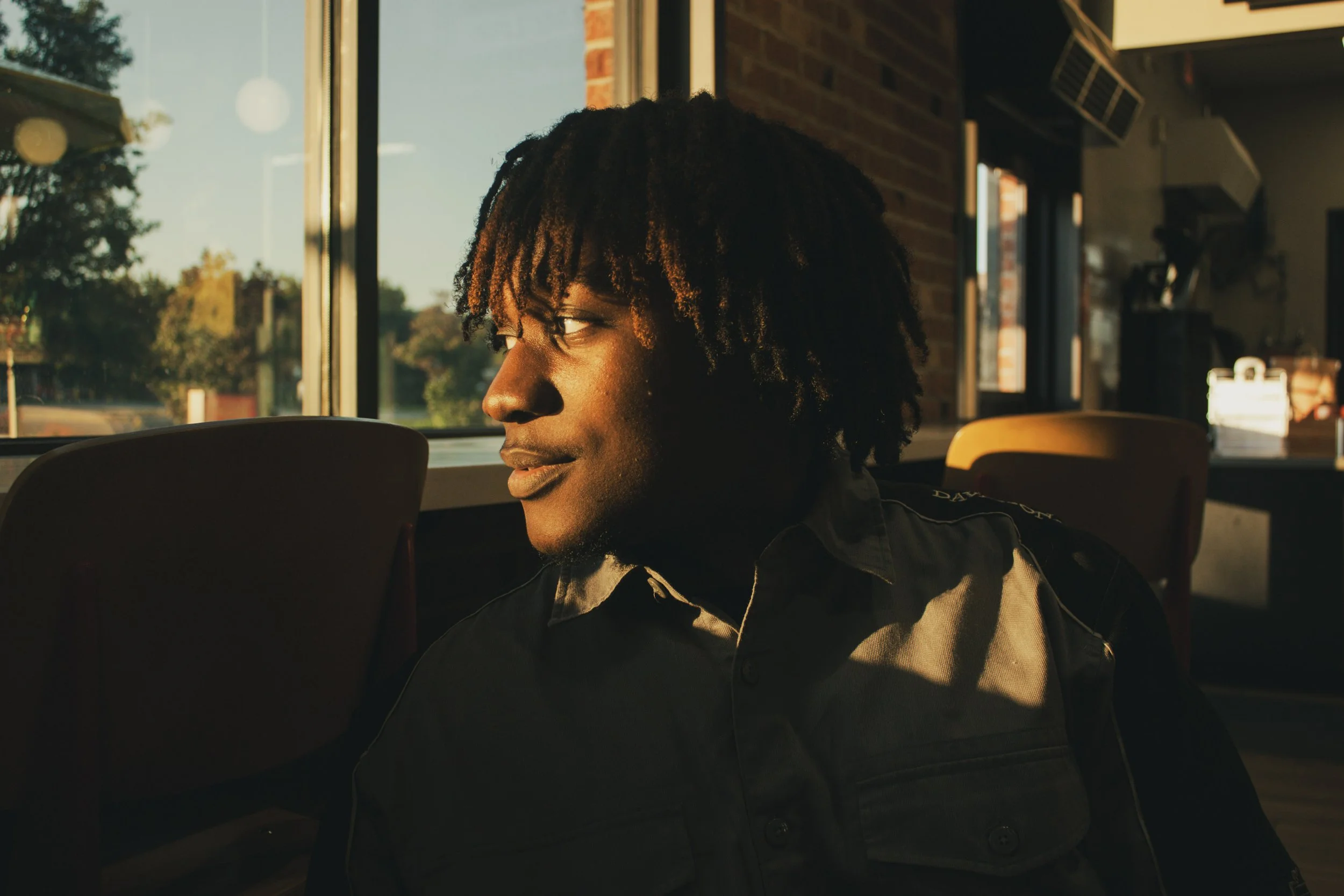 A young man with dreadlocks sitting inside a restaurant, looking towards the window with sunlight illuminating his face.