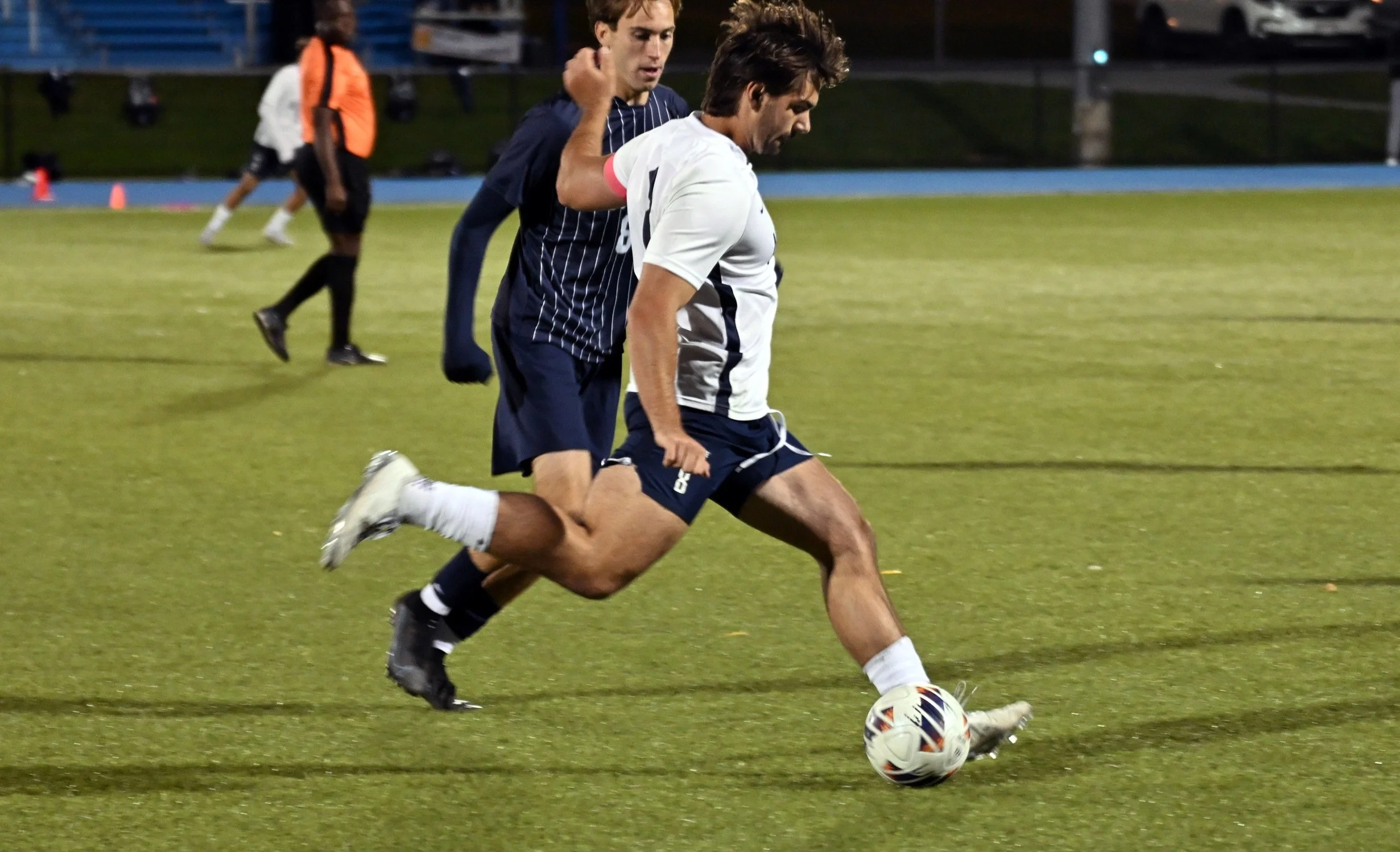 Two male soccer players are competing for control of the soccer ball on a field at night. One player in a white and black uniform is kicking the ball, with a second player in a dark jersey closely defending him. In the background, a person in an oran