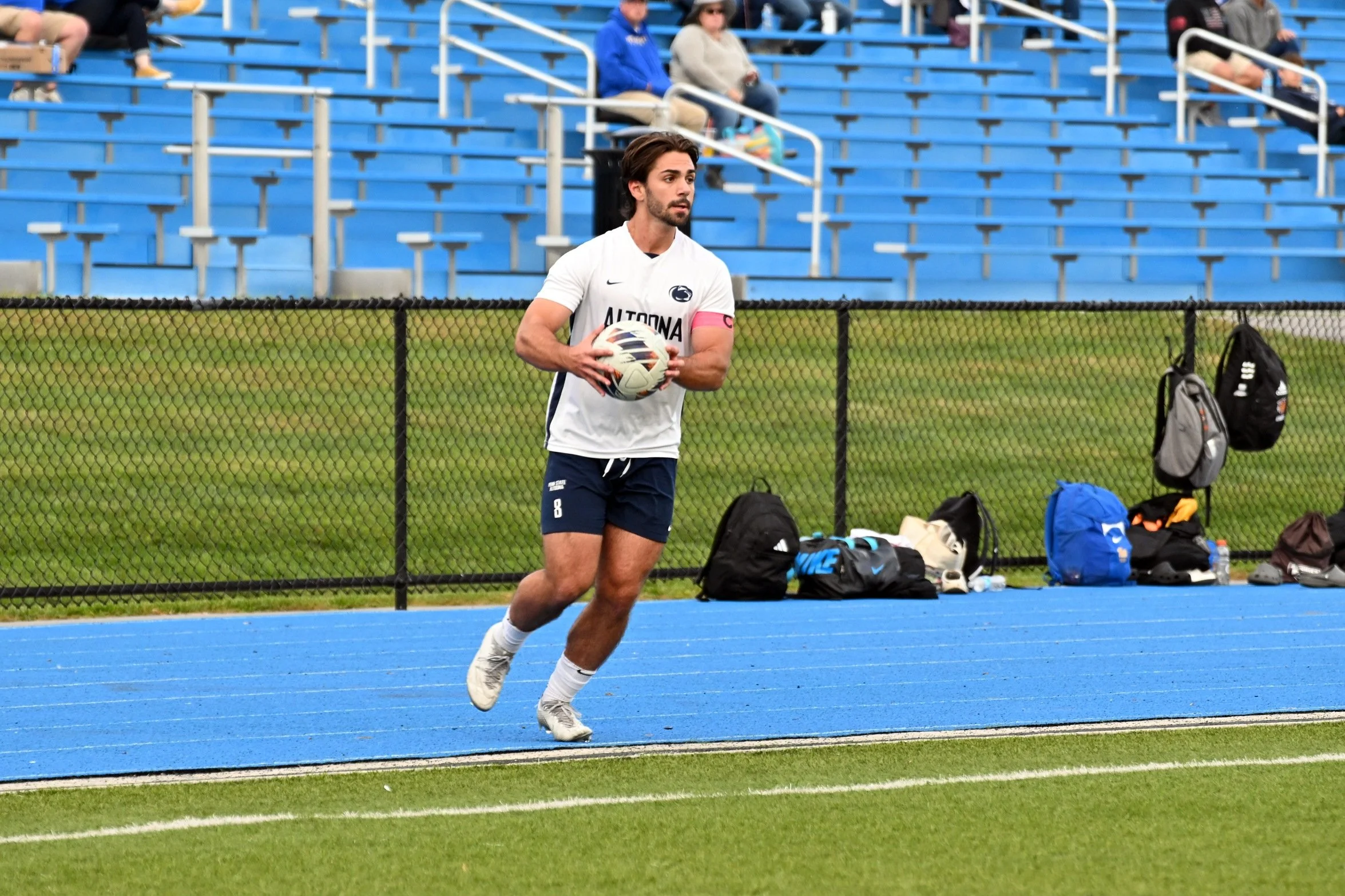 A male soccer player with a beard and brown hair, wearing a white jersey with 'ALTOona' written on it and a pink captain's armband, is holding a soccer ball while standing on a blue running track. In the background, there are blue stadium seats and s