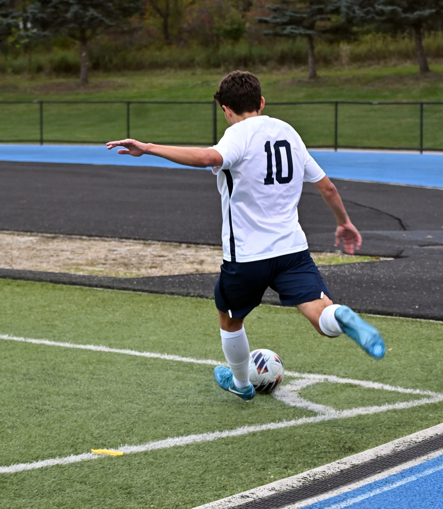 A young male soccer player wearing a white jersey with the number 10, black shorts, and blue shoes is kicking a soccer ball on a grassy field near a running track. He is in motion, with one arm extended and his back facing the camera. In the backgrou