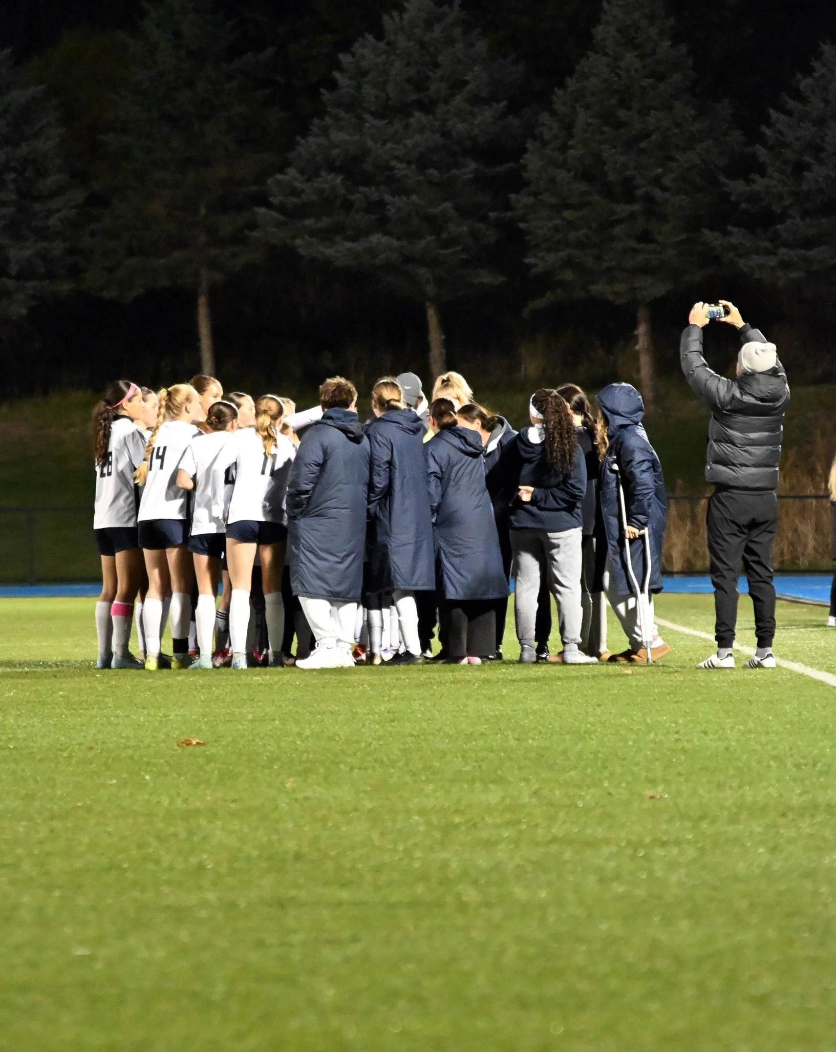 A group of soccer players and coaches gathered on a field at night for a huddle, with some wearing white jerseys and others in dark jackets. A person on the right is taking a photo or video with a smartphone.