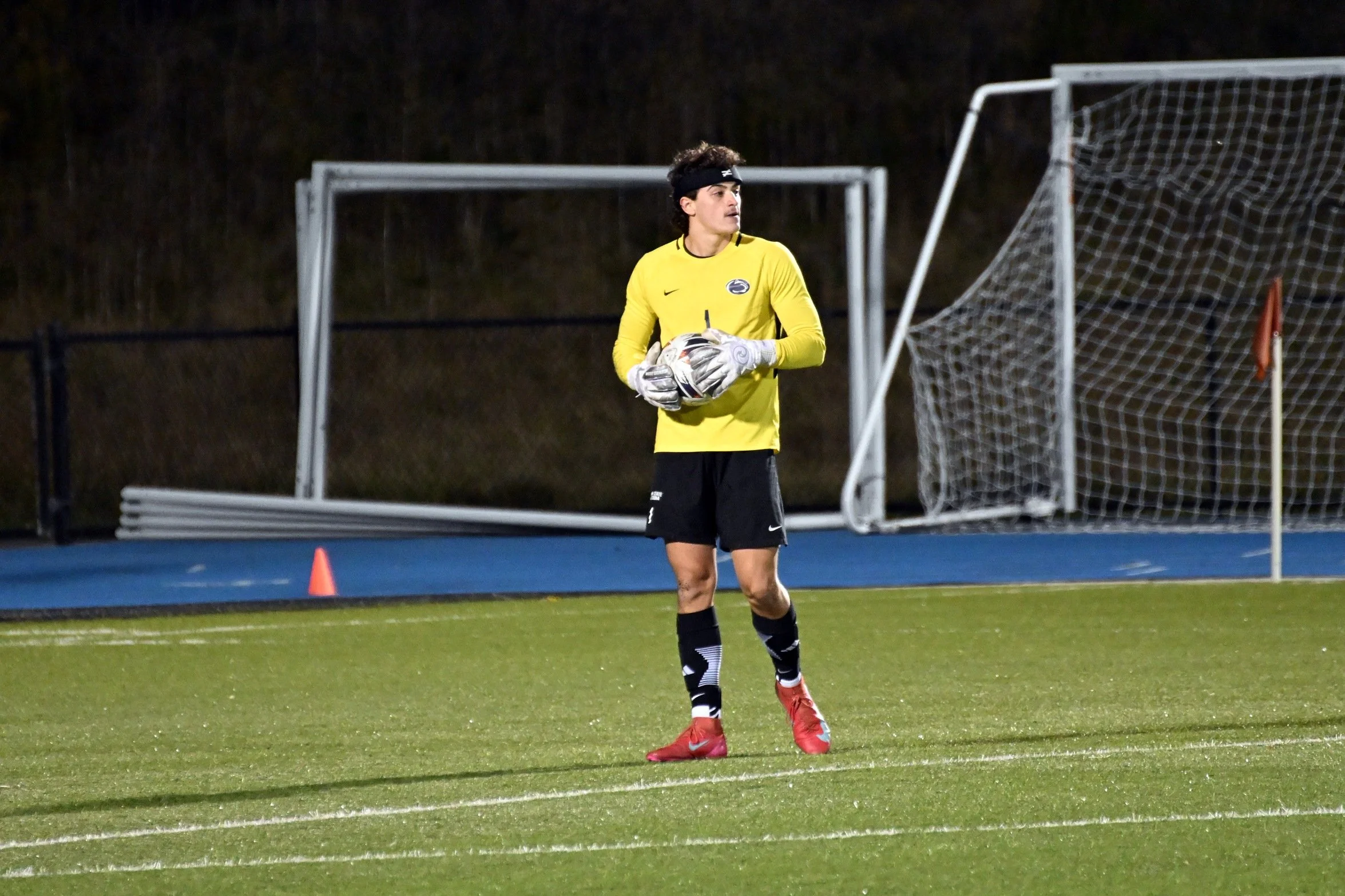 A soccer goalkeeper in a yellow jersey, black shorts, and red shoes holding a soccer ball on a field at night.