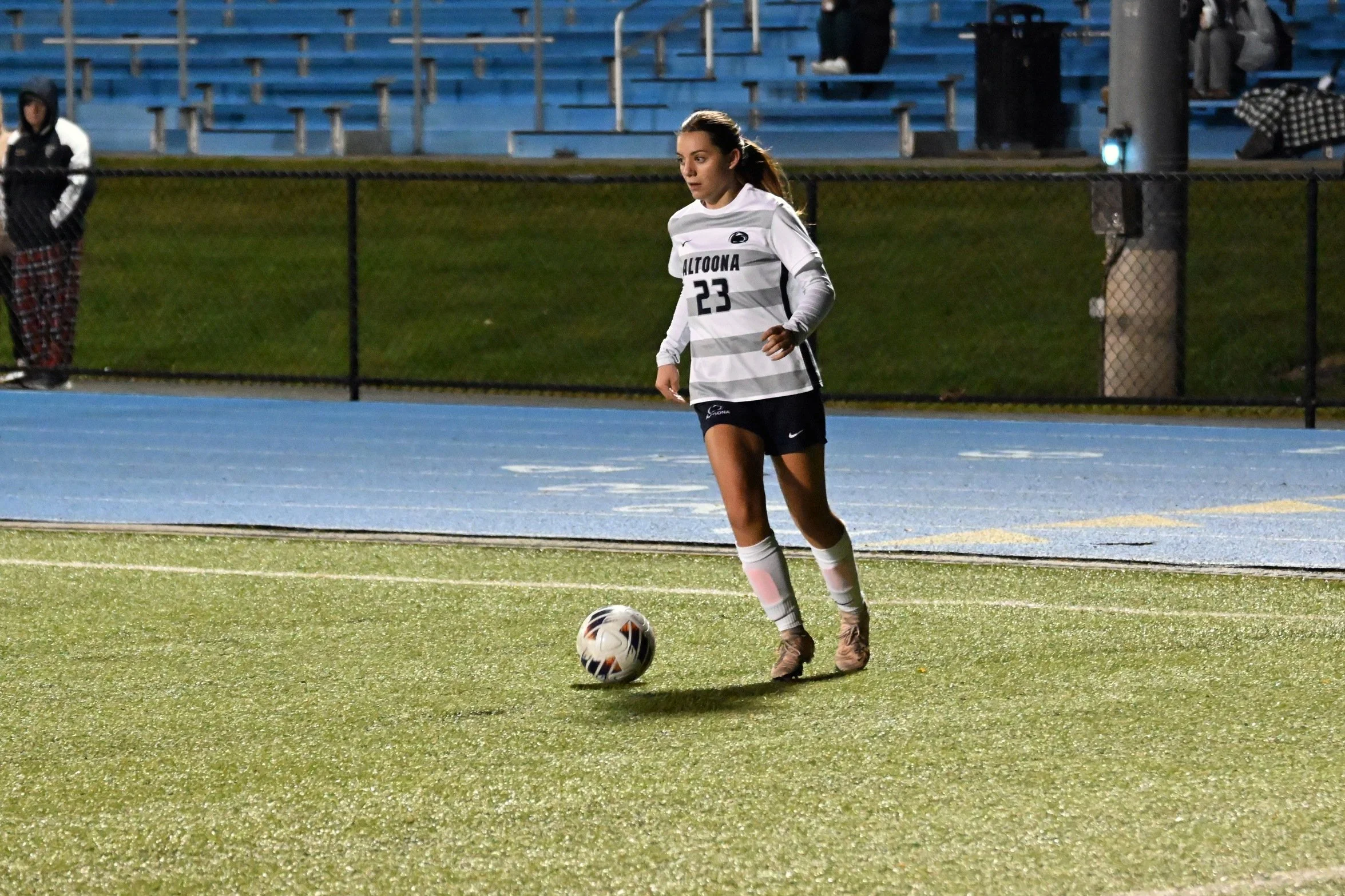 Female soccer player in white and gray striped jersey with number 23 and 'ALTOONA' on front, preparing to kick a soccer ball on a field, during evening or night game.