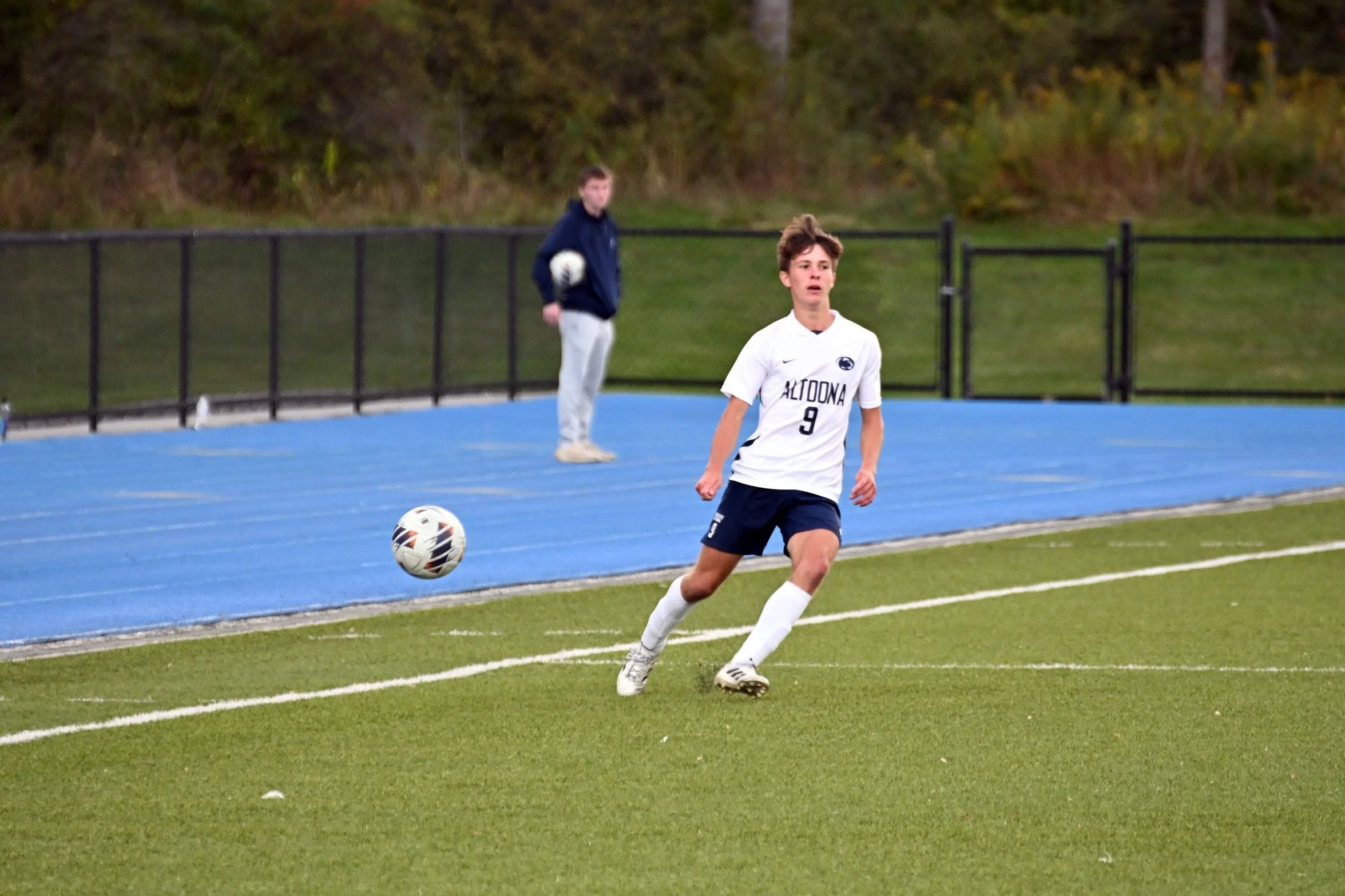 A young male soccer player wearing a white jersey with the number 9 and the word 'ALQUONA' on it, is running on a green soccer field with a blue track in the background. A soccer ball is near his feet, and a person stands behind a black fence holding