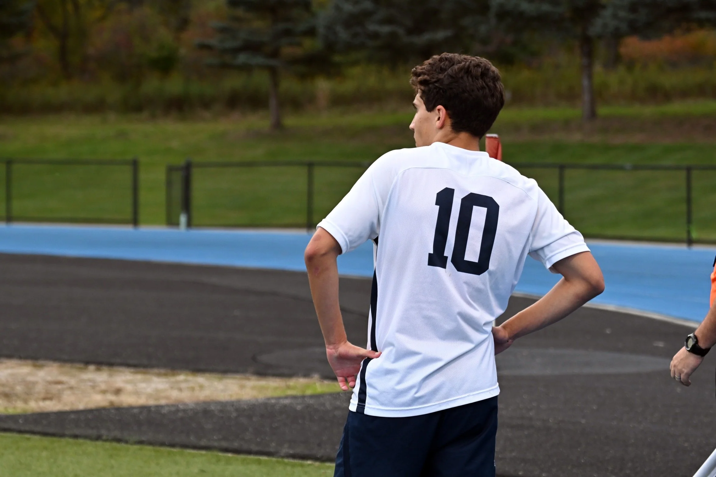 A young man wearing a white sports jersey with the number 10 on the back, standing on a track field during daytime, with his hands on his hips and looking to the side.