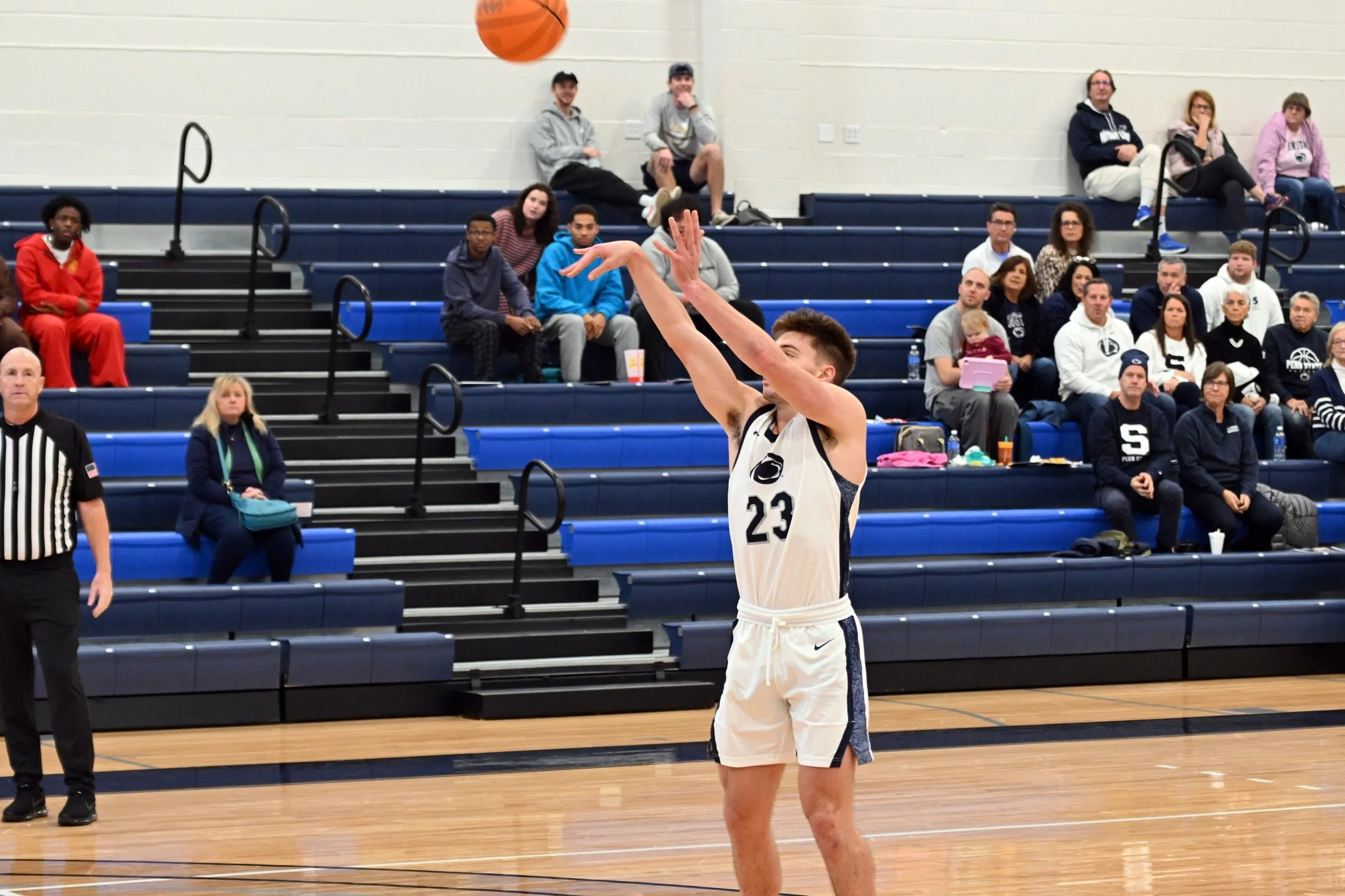 A basketball player in a white uniform with the number 23 is shooting a basketball on an indoor court. Spectators sit on blue bleachers in the background watching the game.