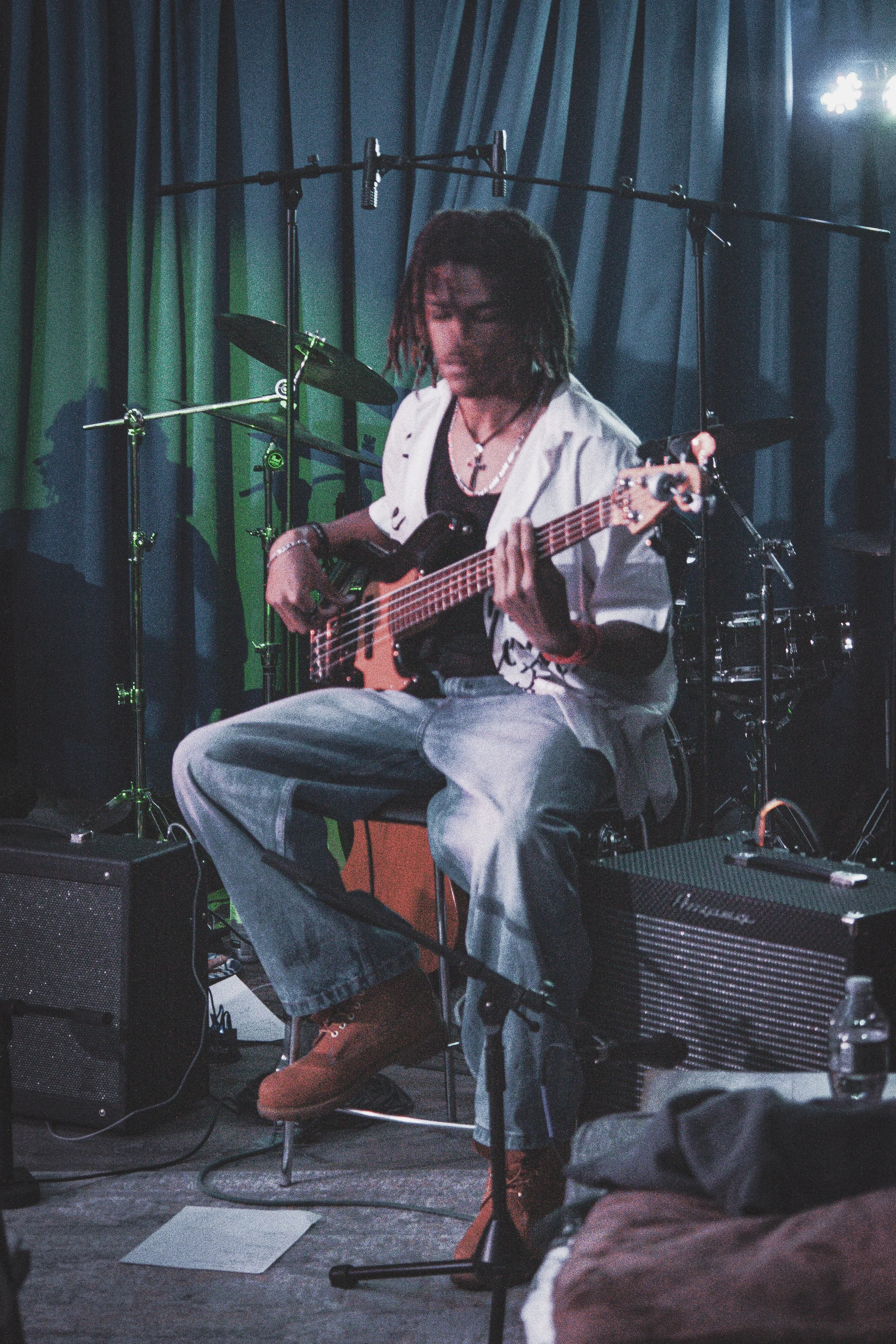 A person playing an electric bass guitar on stage, with drum set and music equipment around, illuminated by stage lights.