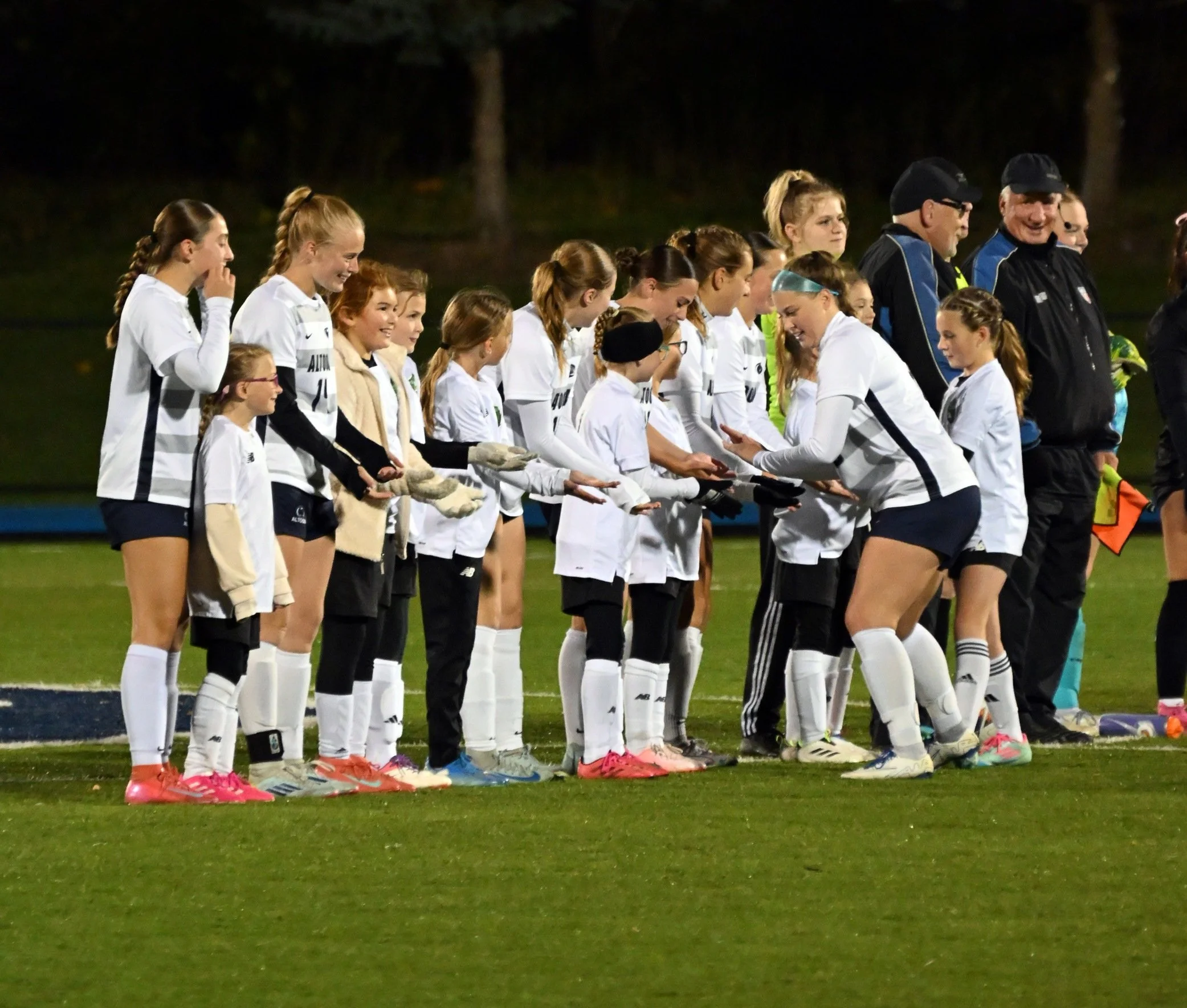 A group of young female soccer players and their coaches standing in a line on a soccer field at night, wearing sports uniforms and smiling.