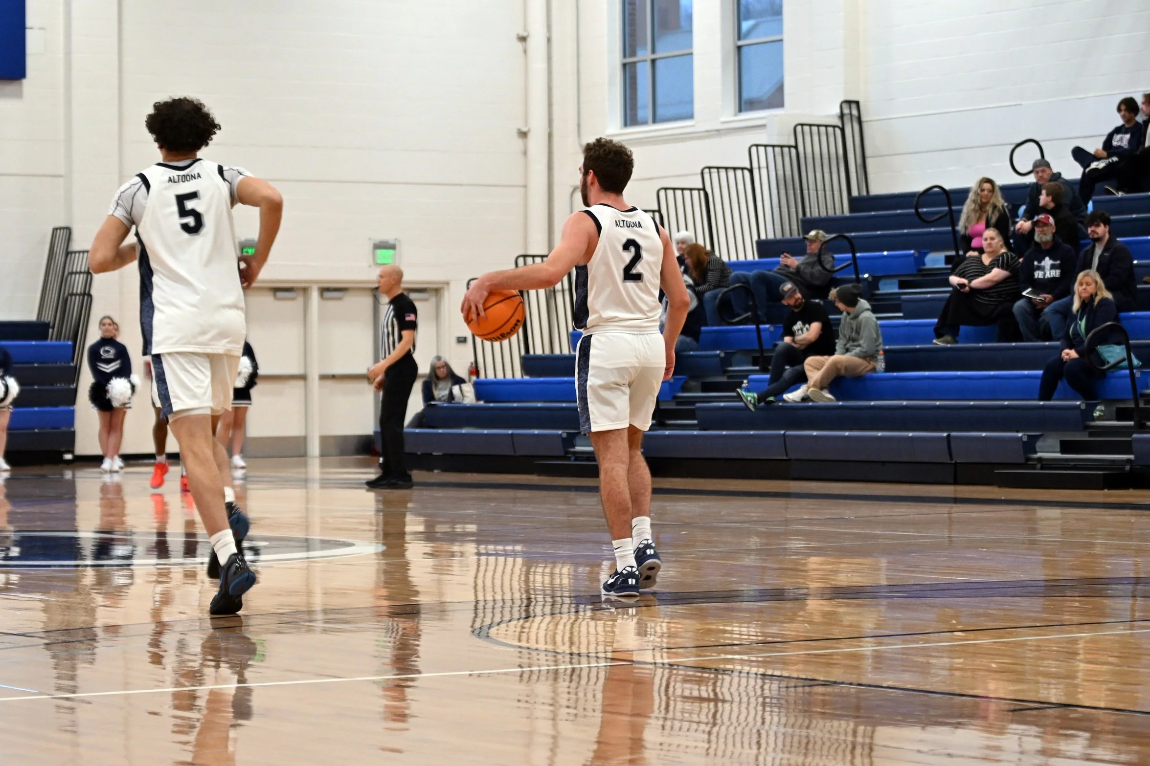 Two basketball players in white uniforms with black trim, numbered 2 and 5, on a wooden indoor court. One is holding a basketball, and there are spectators seated in the blue bleachers in the background.