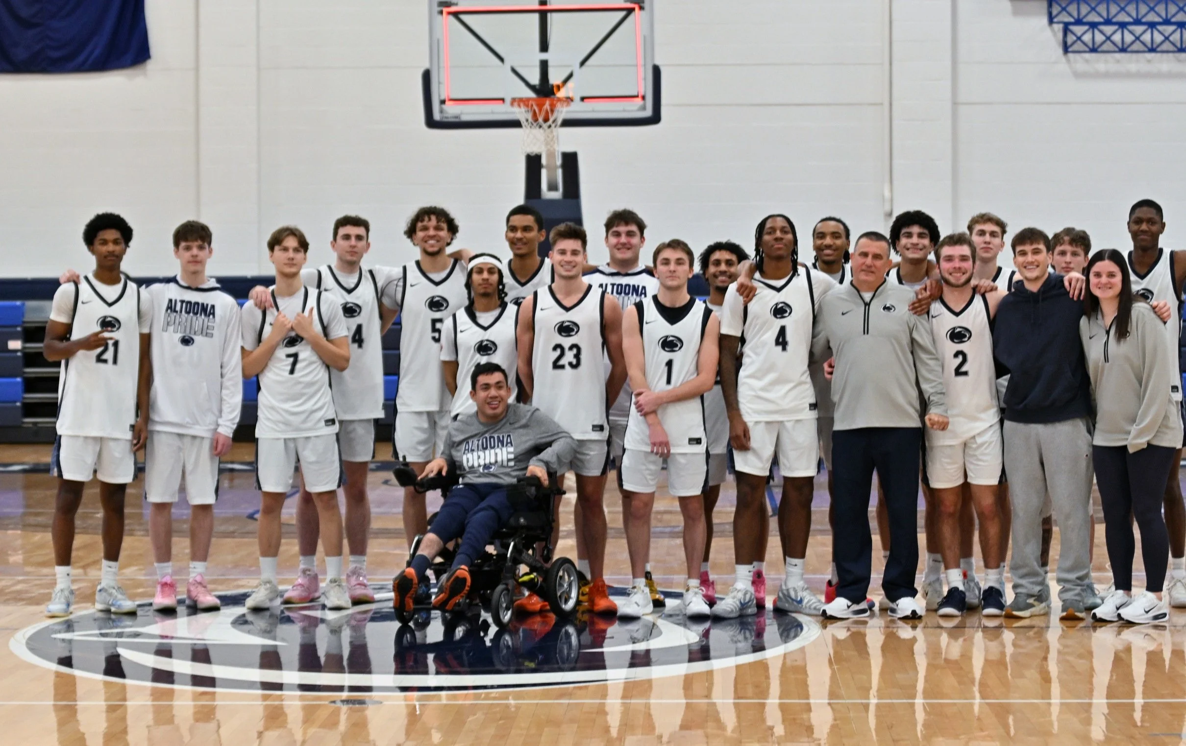 A diverse group of young men and women, including some with darker skin, stand together on a basketball court in an indoor gymnasium, wearing sports jerseys, with one person in a wheelchair at the front. They are smiling and posing for a team photo, 