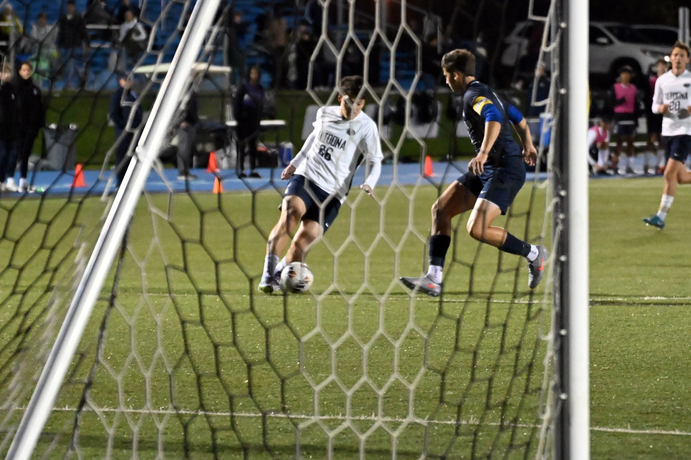 Two soccer players are near the goal post, competing for the ball during a match, with spectators watching in the background.
