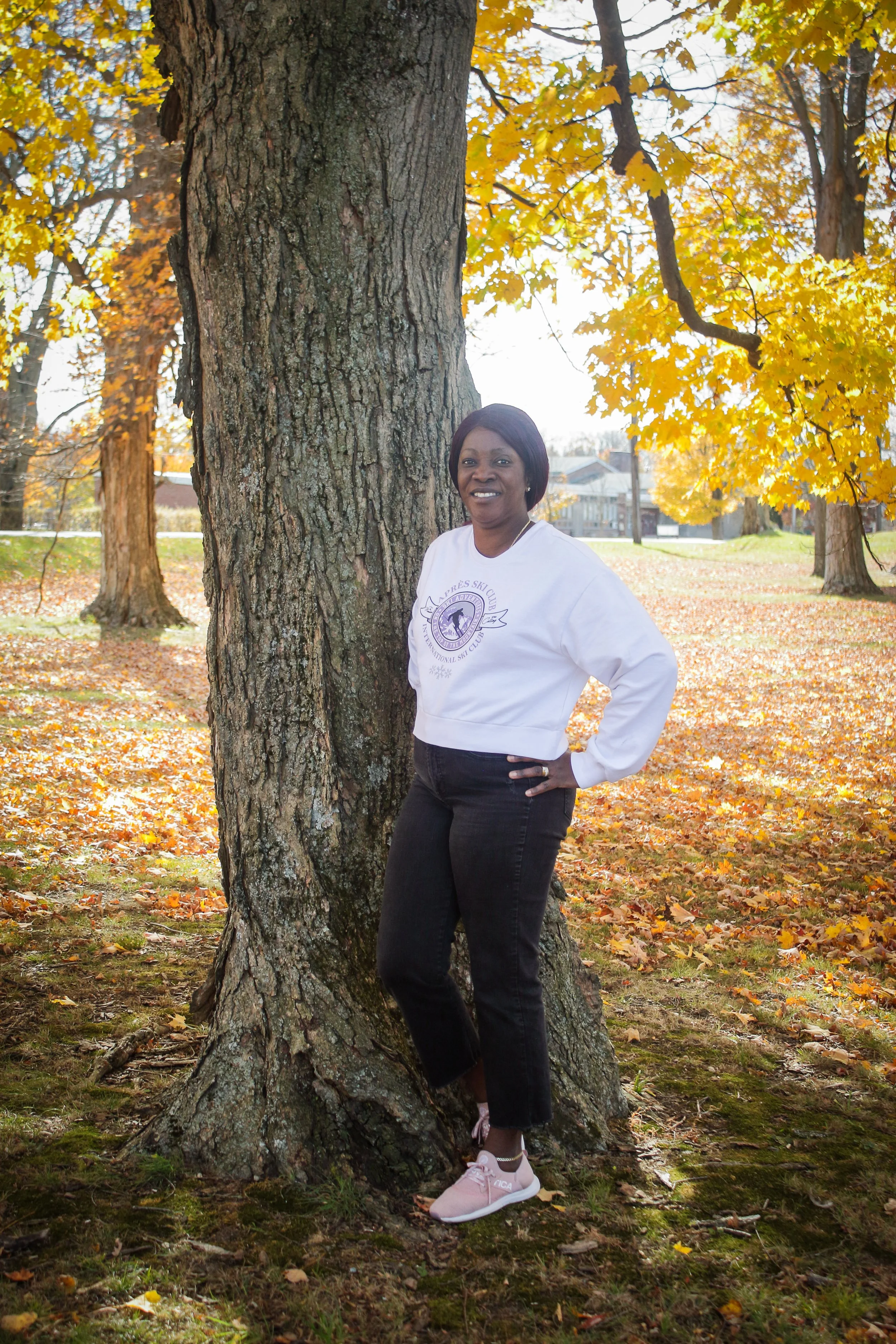 A woman stands outdoors beside a large tree with yellow autumn leaves. She is smiling, wearing a white sweatshirt with a logo, black jeans, and pink sneakers. The background shows more trees with fall foliage and a grassy area scattered with fallen l