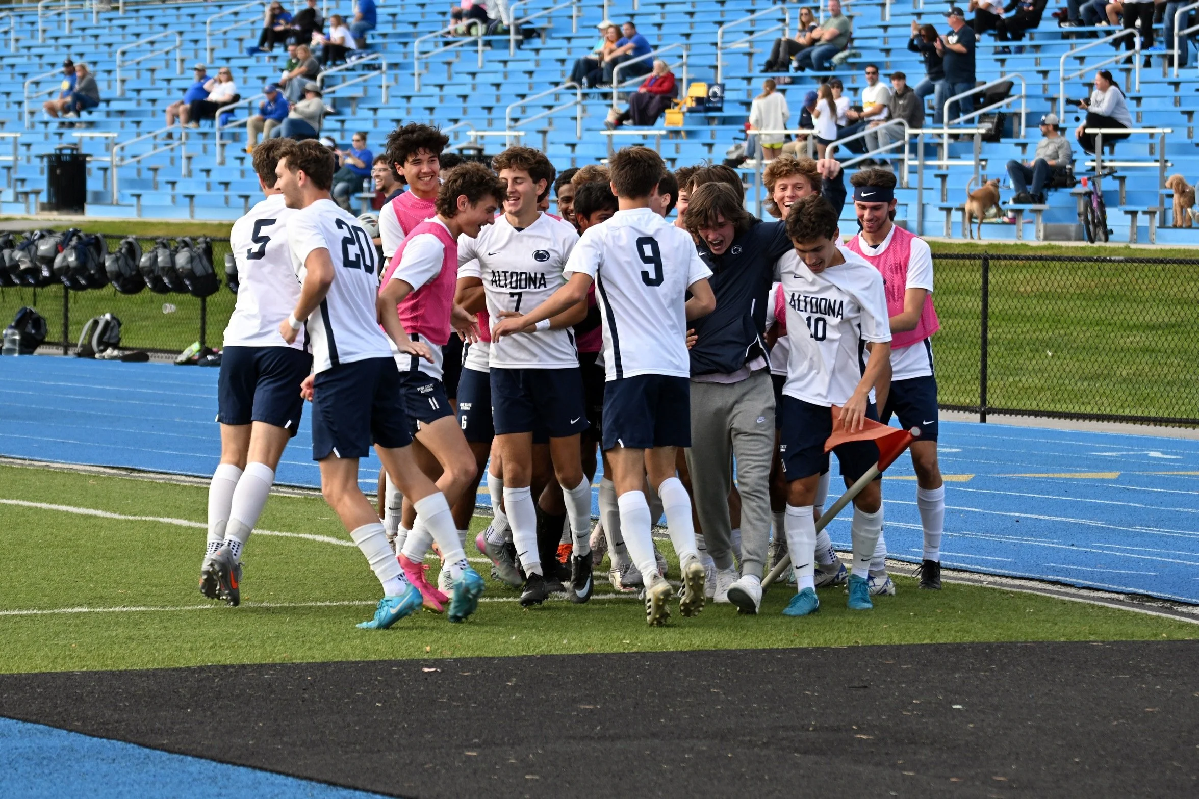 A group of young male soccer players celebrating together on the field, wearing white and pink jerseys, with blue stadium seats and spectators in the background.
