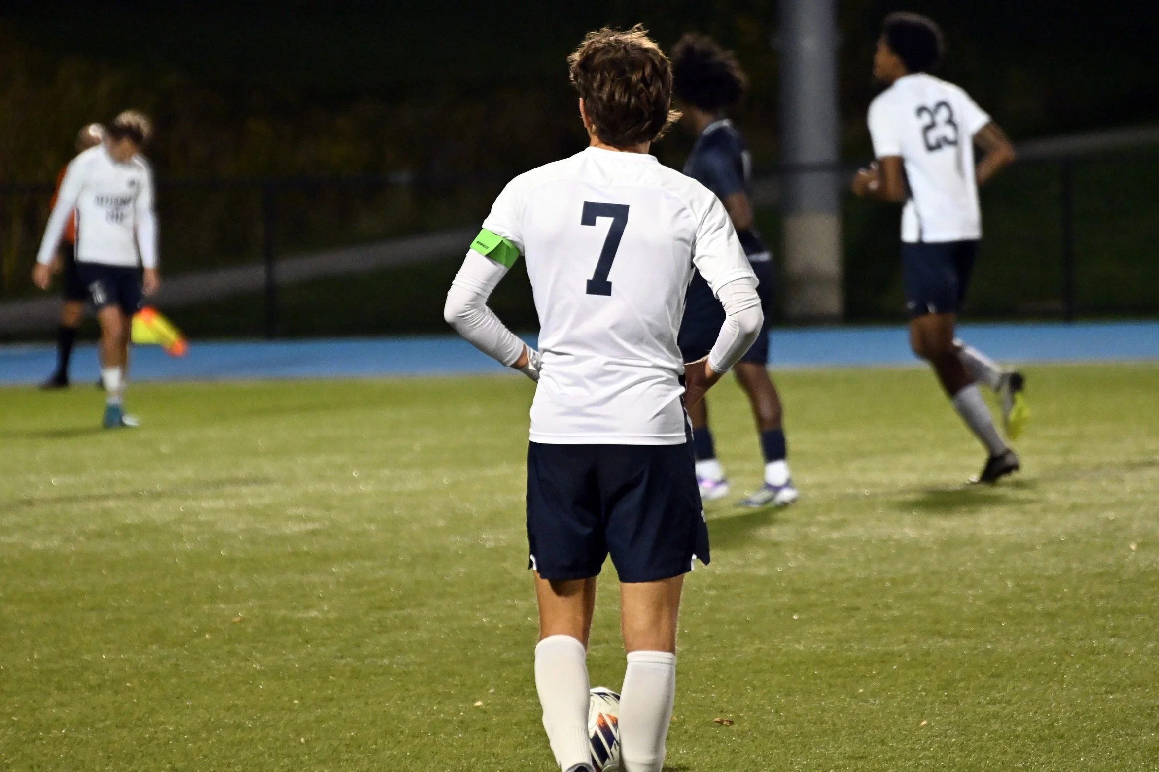 A soccer player wearing a white jersey with the number 7 and a captain's armband, standing on a field at night with other players visible in the background.
