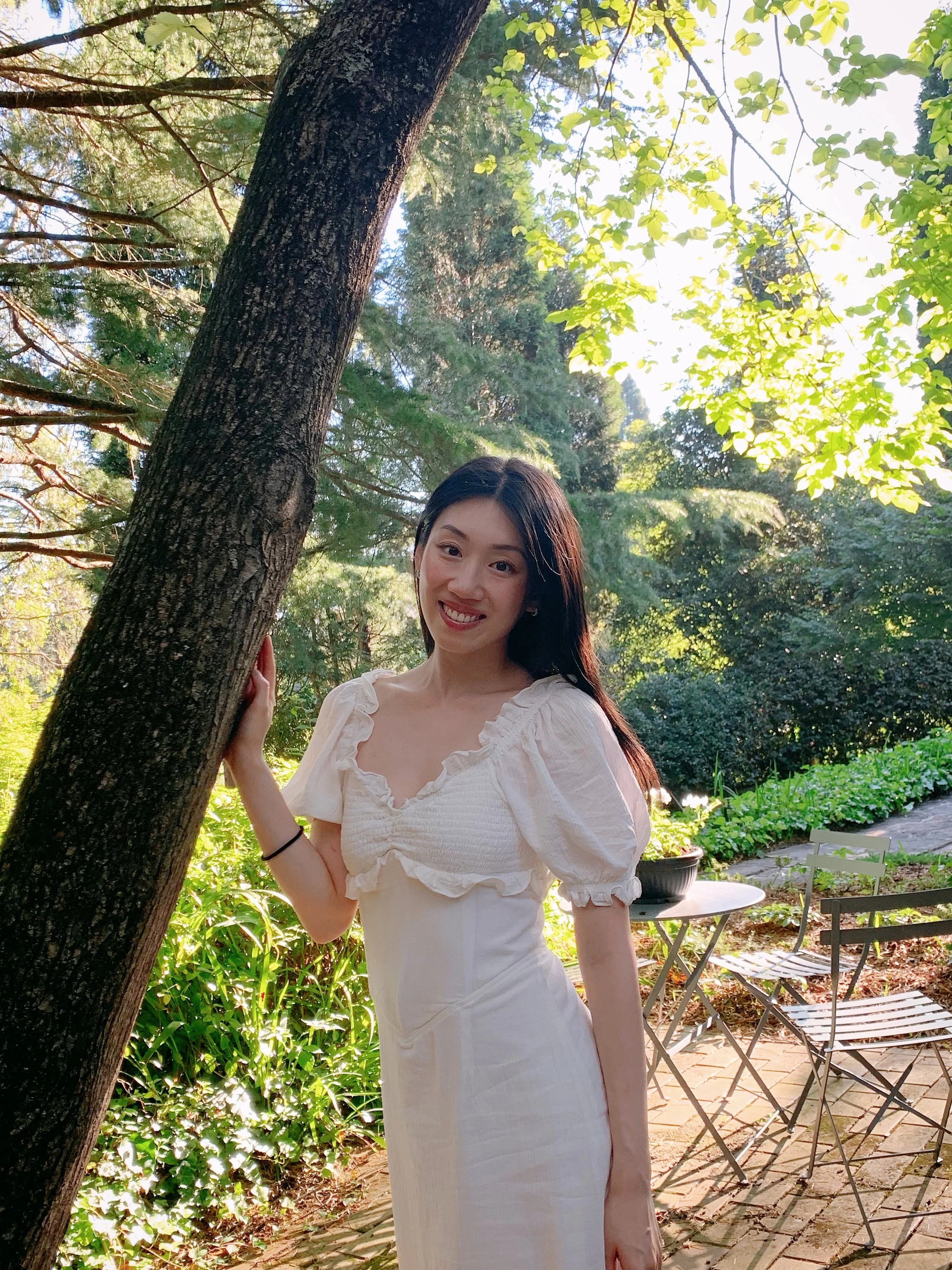 A woman in a white dress smiling while standing outdoors among trees and greenery, holding onto a tree trunk.