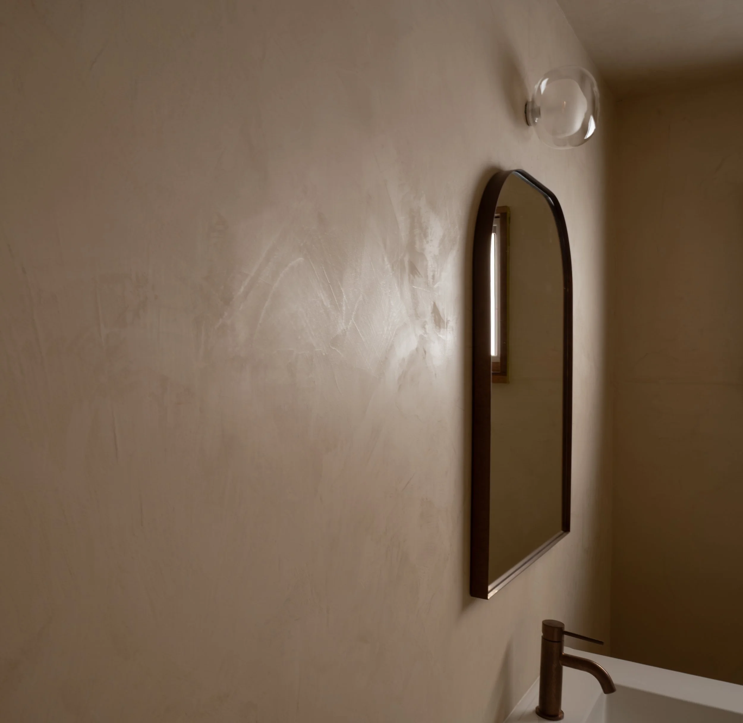 A bathroom sinks area with a copper faucet, a large mirror, a clear glass globe light fixture on the ceiling, and beige walls.