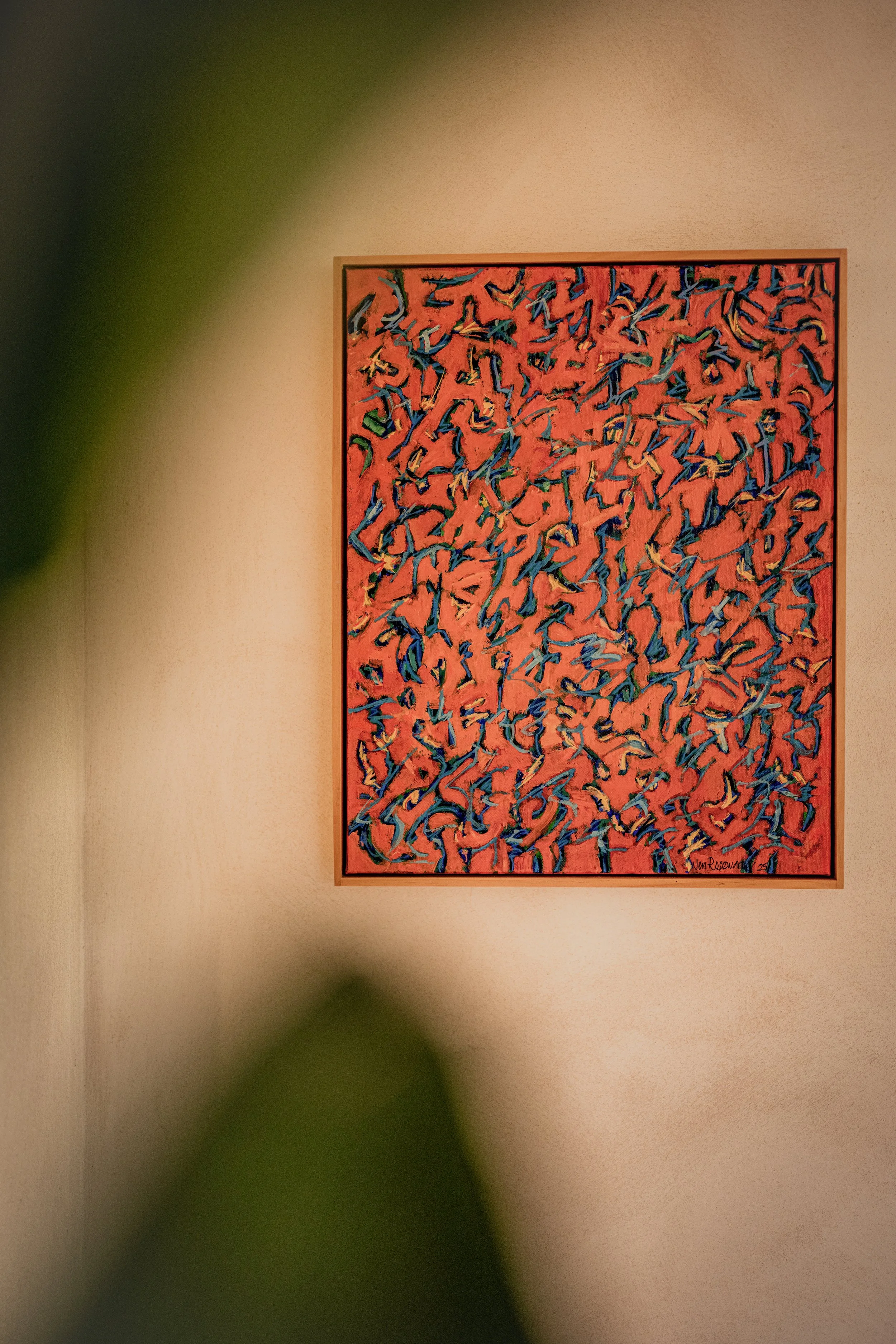 An abstract painting with dynamic, swirling blue, green, and yellow strokes on a pink background, framed and hung on a beige wall.