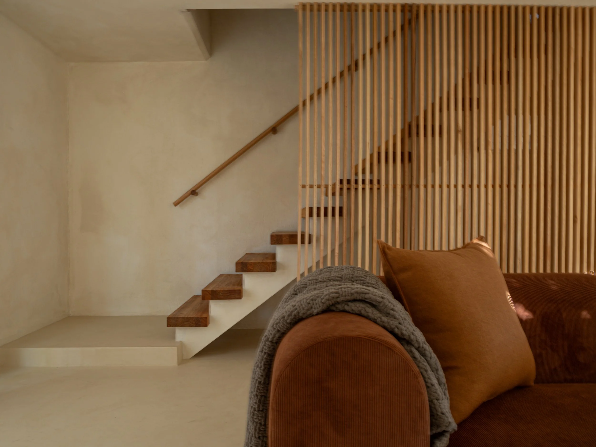 Interior view of a staircase with wooden steps and a wooden slat railing. Part of a brown sofa with a gray throw blanket is visible in the foreground.