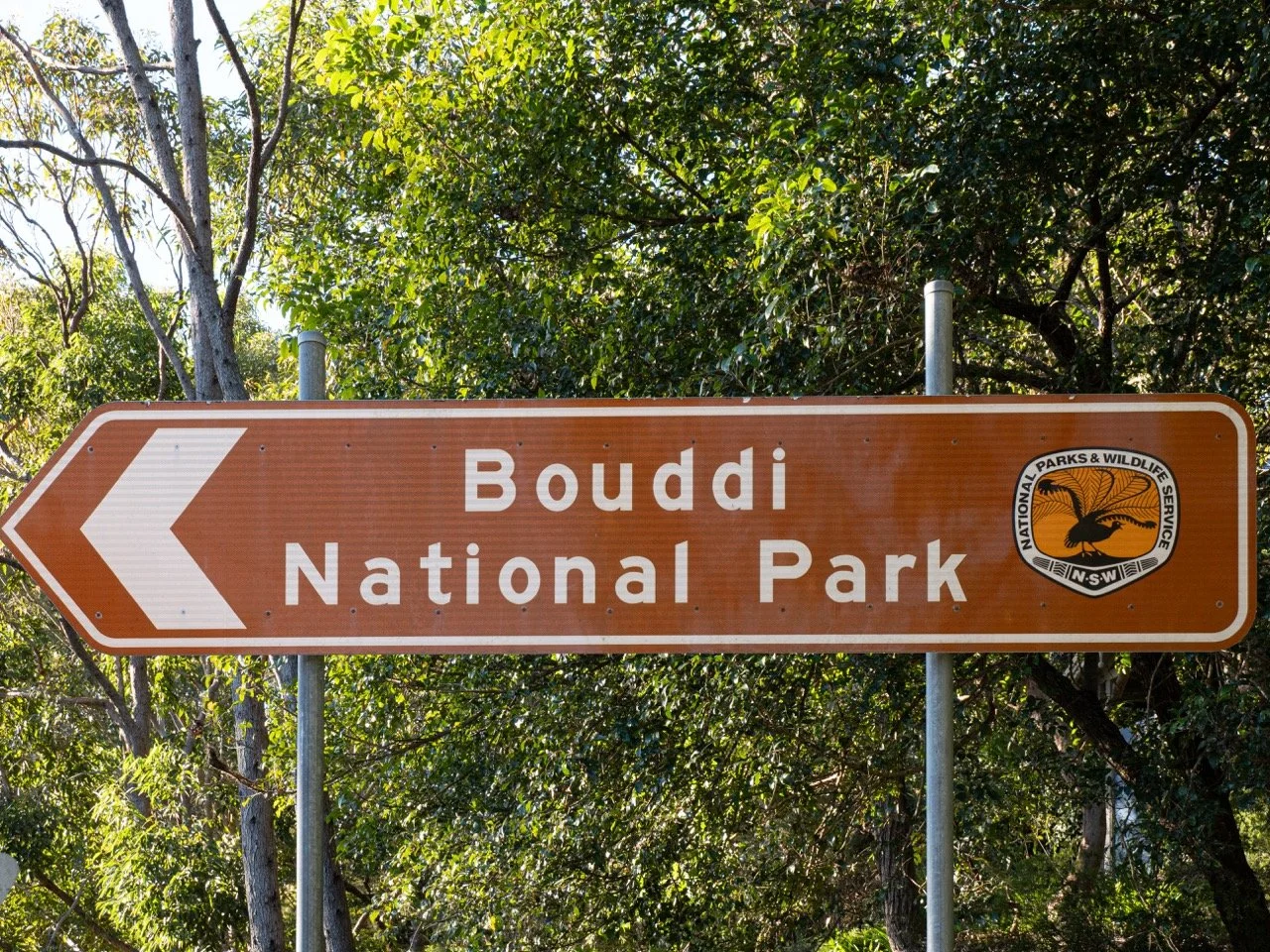 Brown directional sign for Bouddi National Park, pointing left, with the Parks & Wildlife Service emblem on the right, facing trees and greenery.