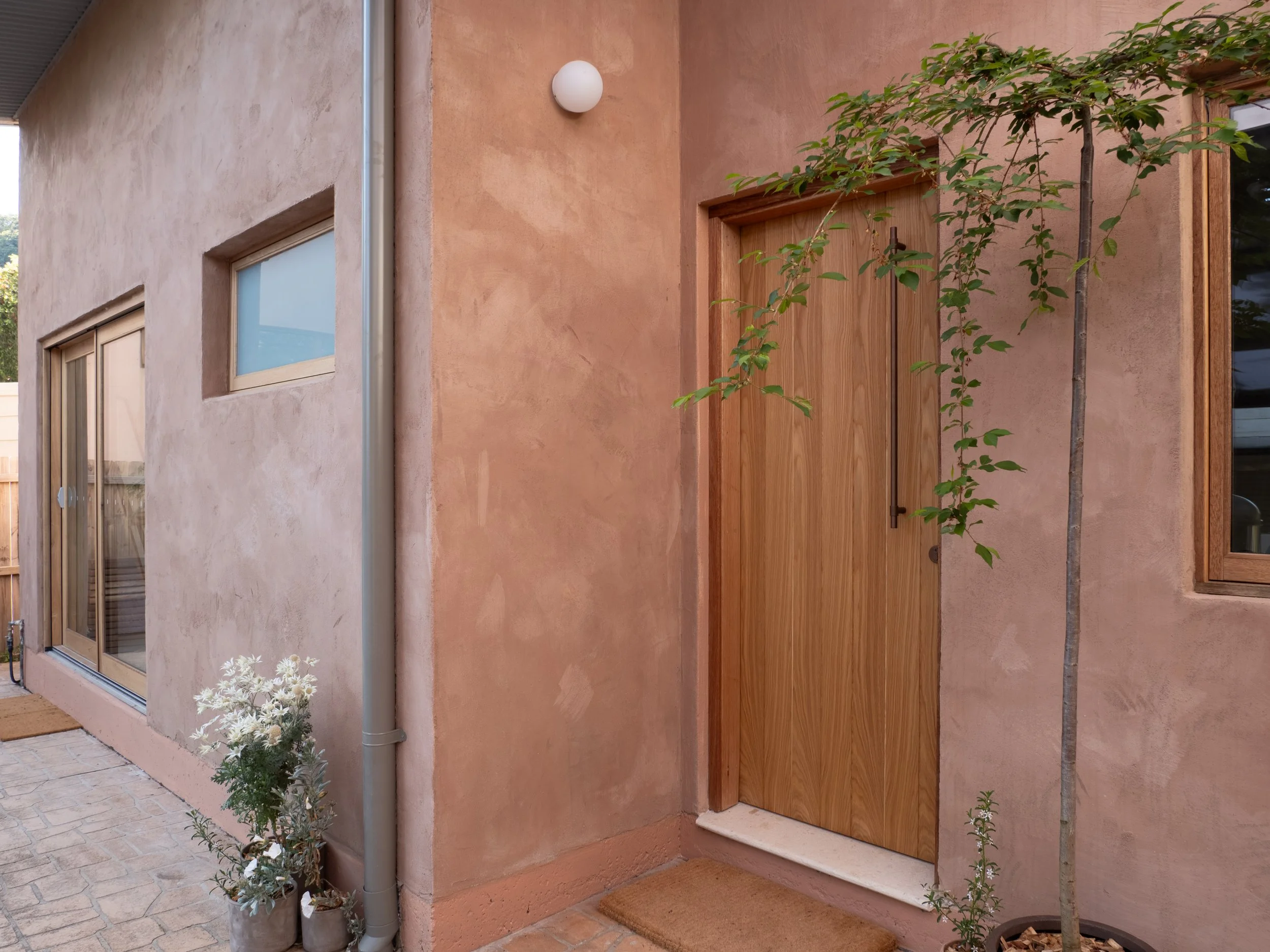 The front entrance of a house with a wooden door, pink stucco exterior wall, small trees, and flowering plants.