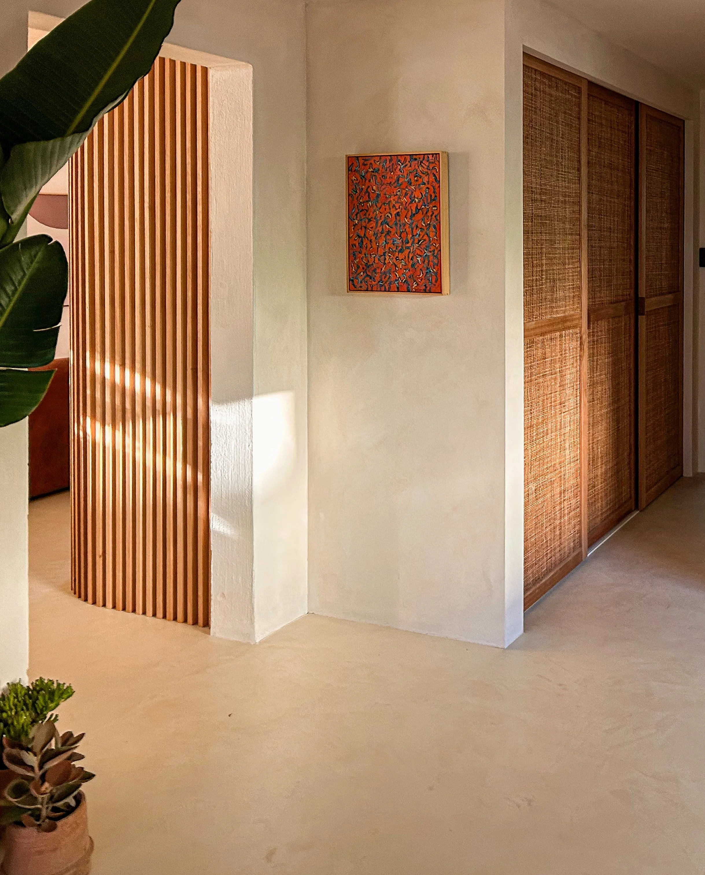 Interior view of a room with a beige floor, white walls, a wooden slatted partition on the left, a red abstract painting on the wall, and wooden sliding closet doors on the right. A potted plant with broad green leaves is partially visible in the cor