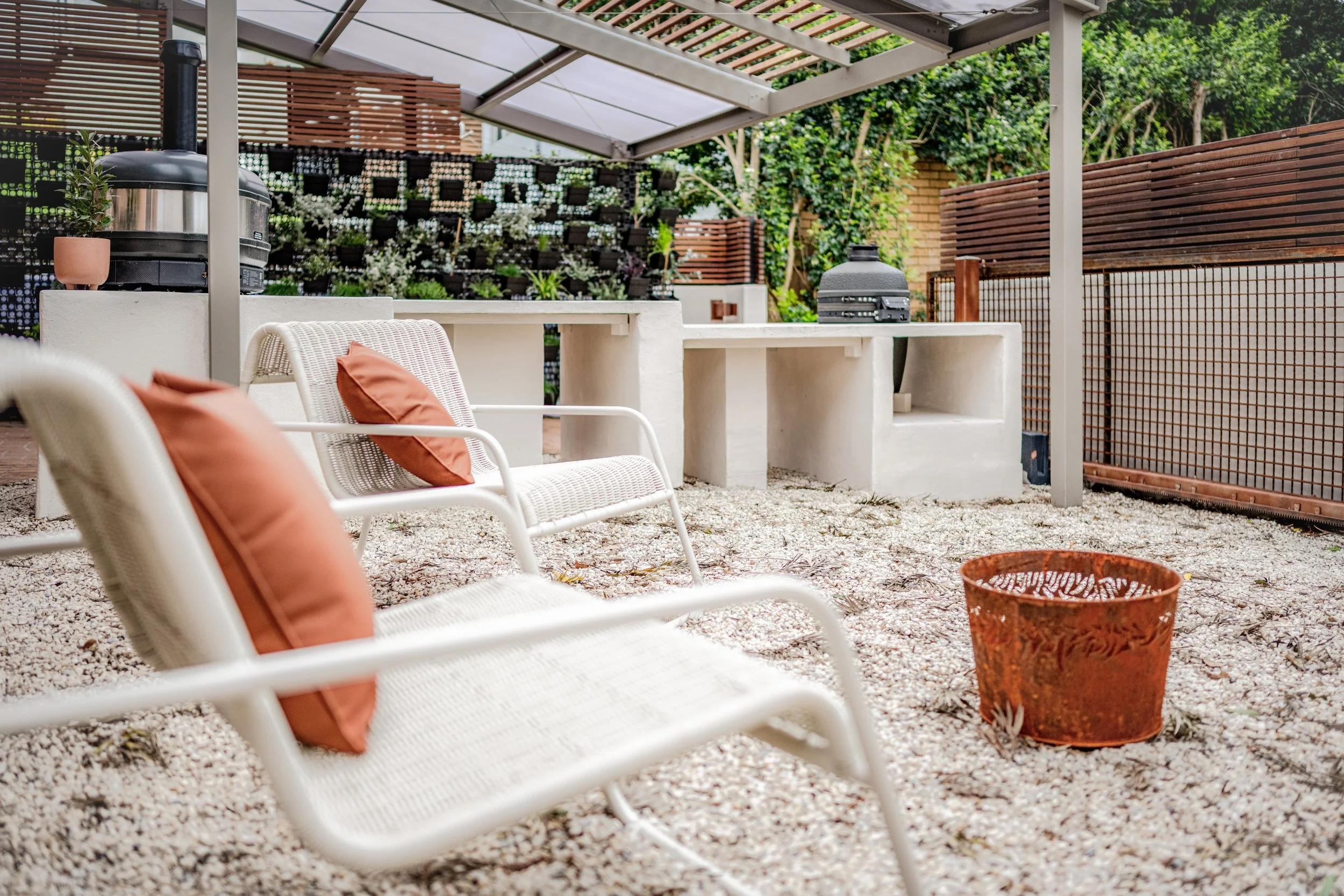Outdoor patio area with white chairs and orange cushions, gravel ground, a small copper-colored decorative vase, and a covered grill station with potted plants and a vertical garden in the background.