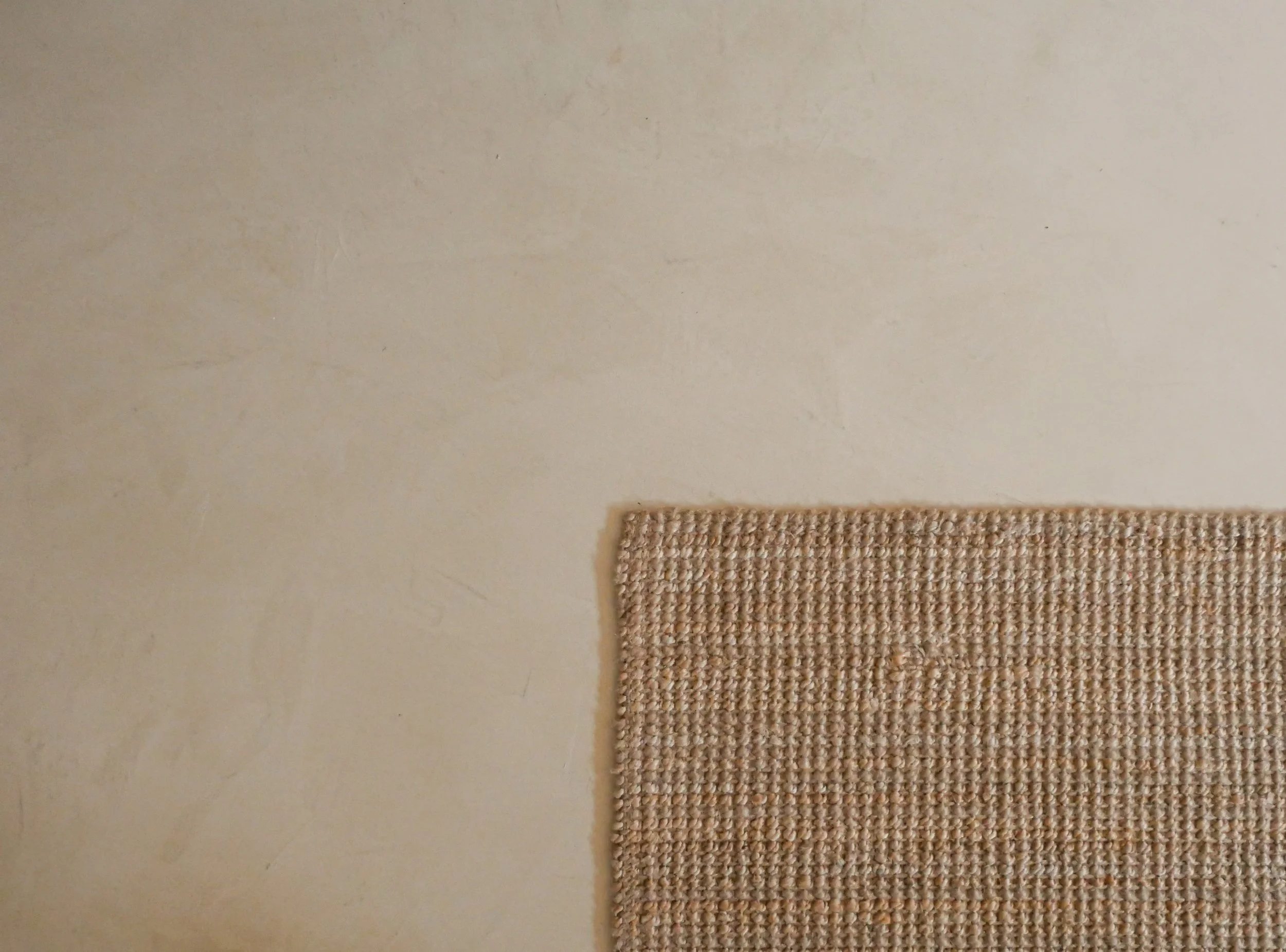 Close-up of a beige woven textile next to a slightly rough painted wall in a light beige or cream color.