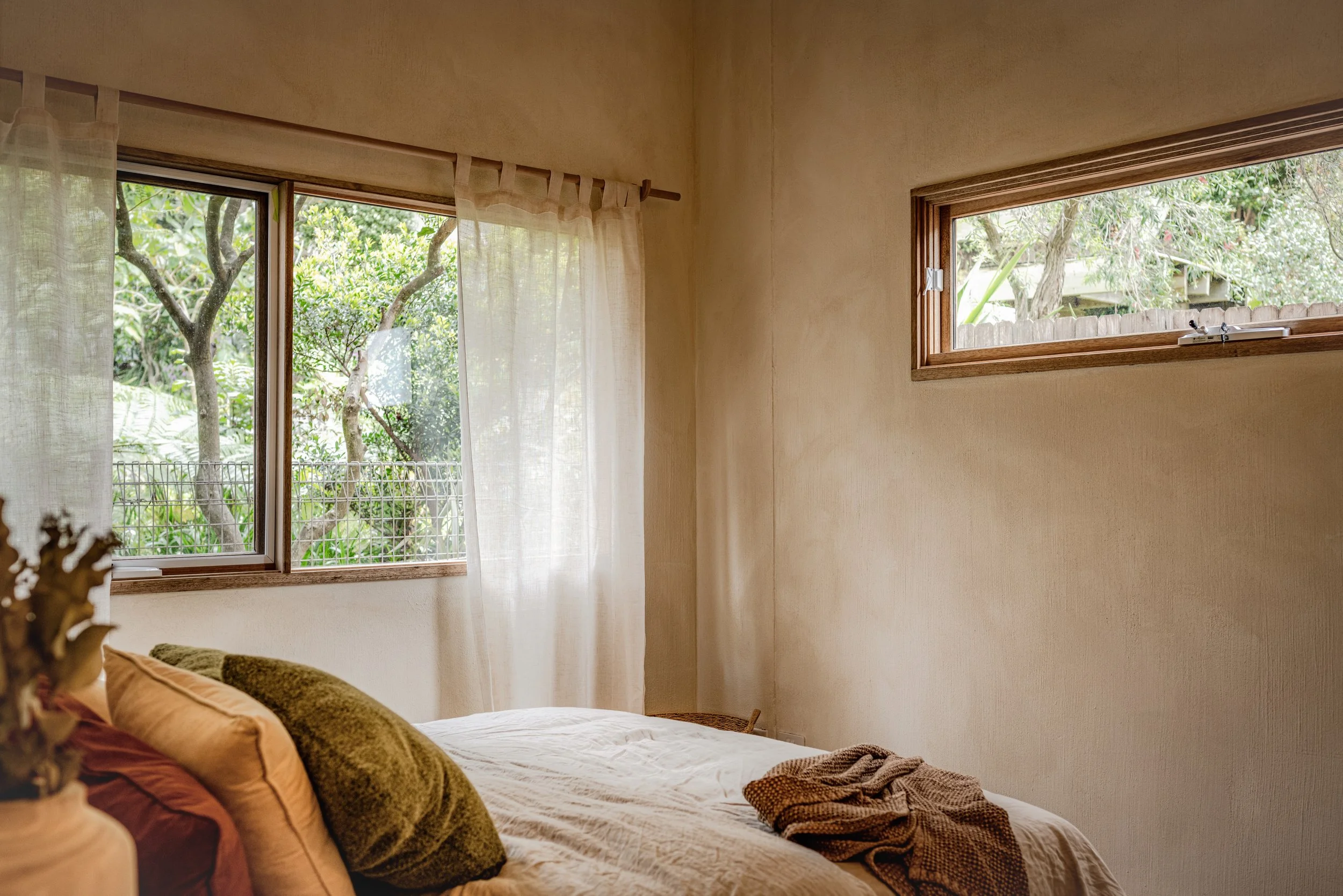 A bedroom with two windows showing green trees outside, light-colored walls, a bed with pillows and a blanket.