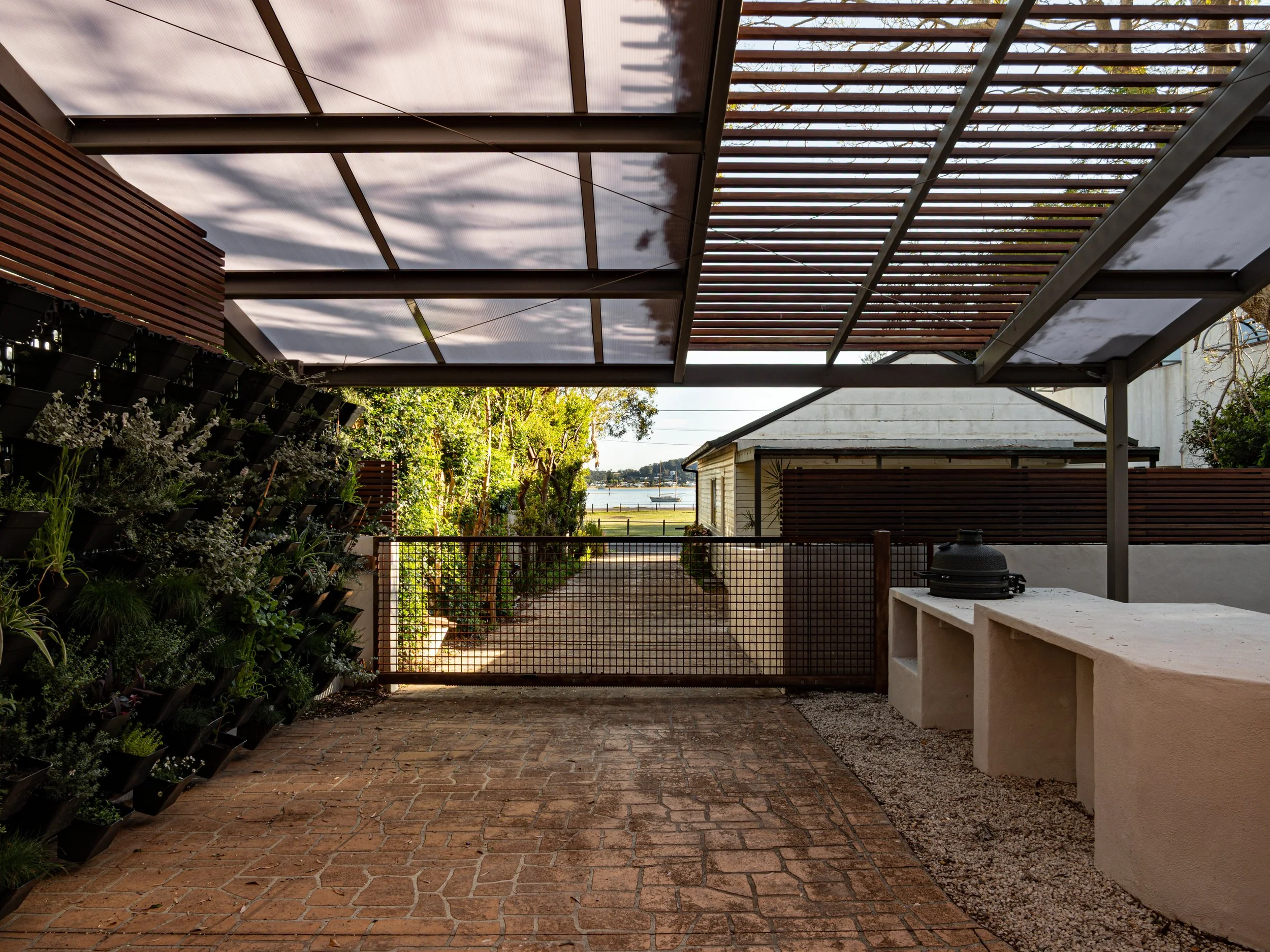 Backyard with brick patio, green plants, a wooden fence, and a lattice roof structure with sunlight filtering through.