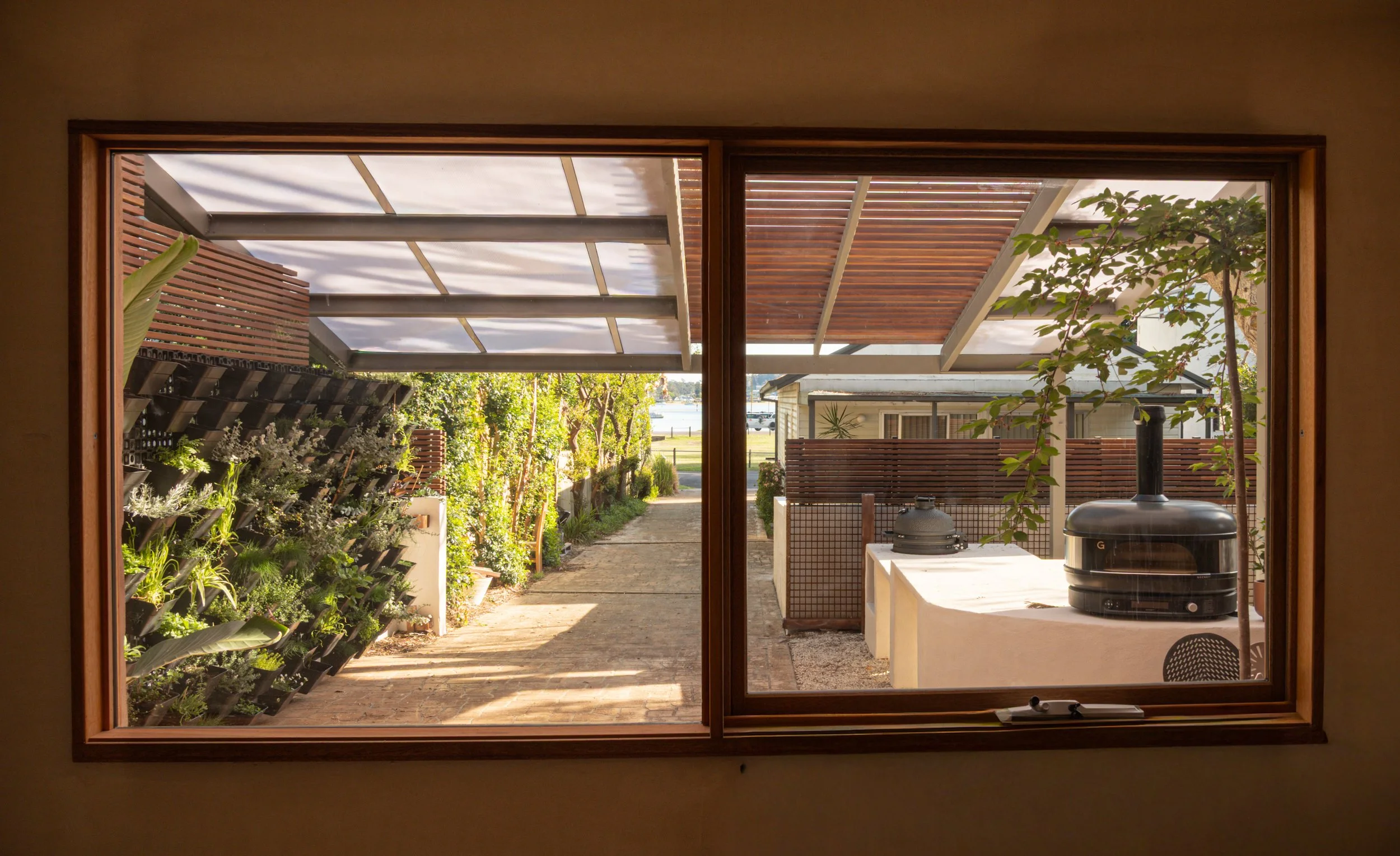View through a large glass window of a backyard garden with a pathway, plants, and a shade structure overhead. There is a barbecue grill on a stone counter inside the house near the window.