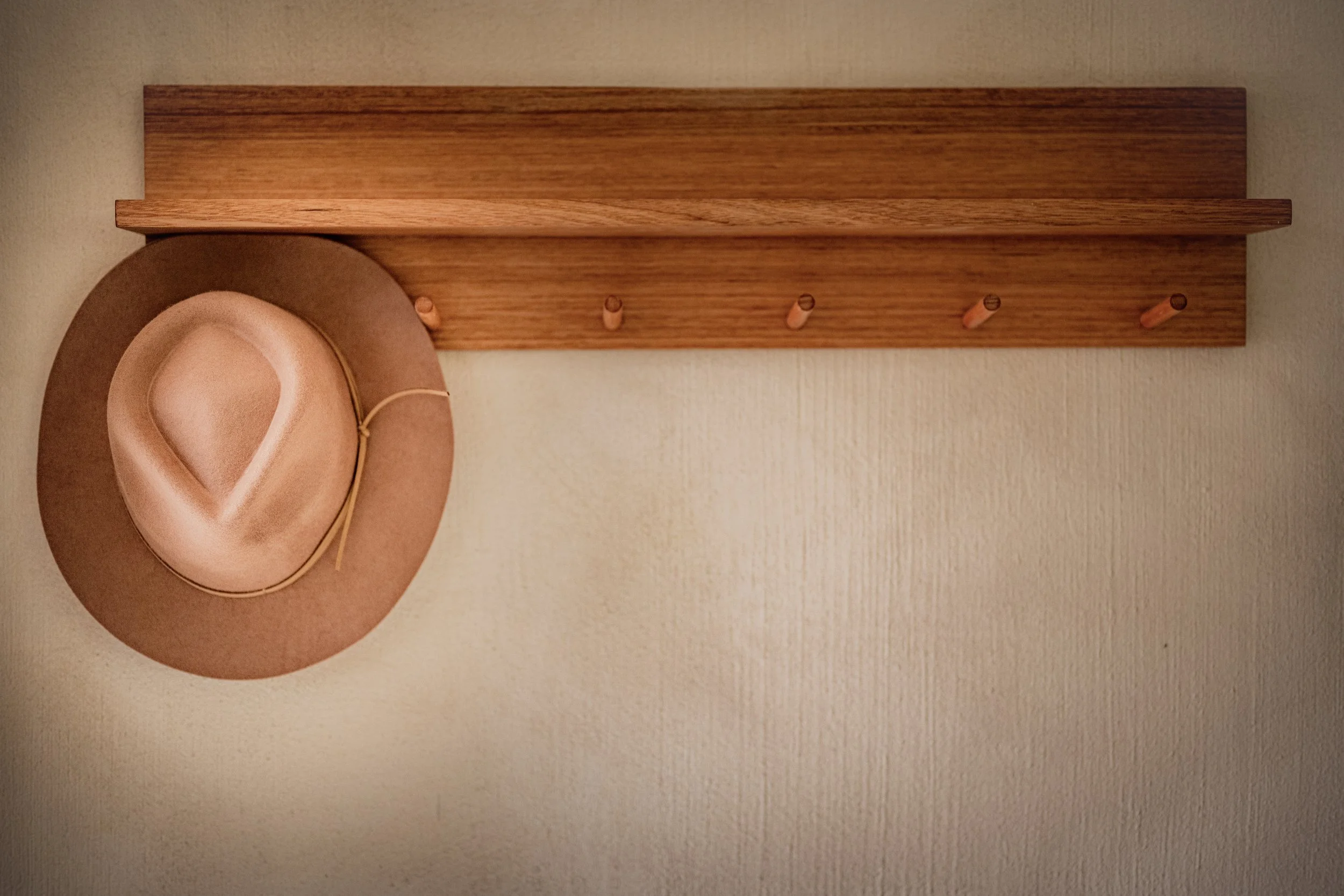 A wooden wall-mounted coat rack with four hooks, next to a beige hat hanging on one of the hooks.