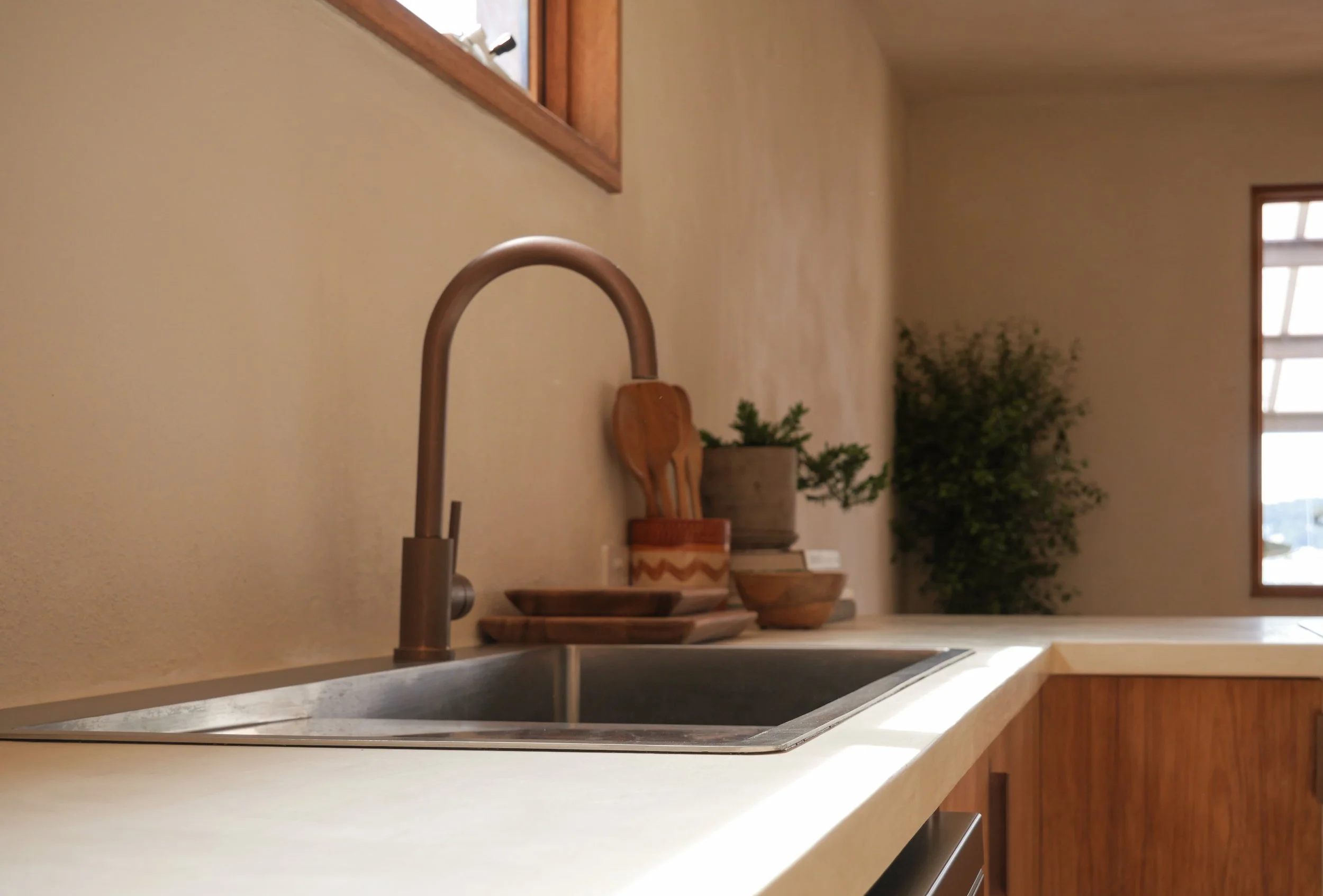 Close-up view of a kitchen countertop with a copper faucet, wooden bowls, and decorative green plants against a beige wall, with a window in the background.