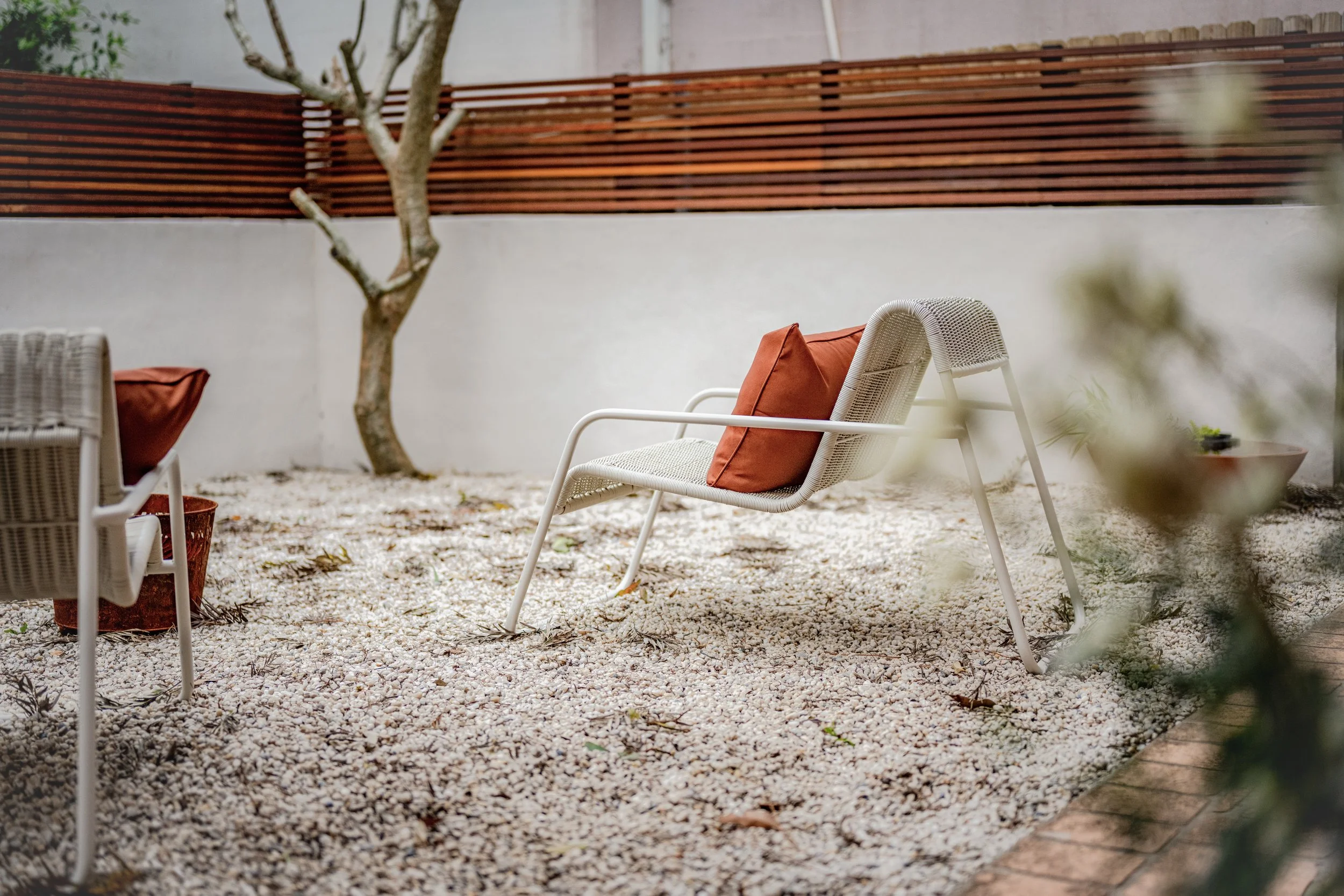 A minimalist outdoor patio with a white lounge chair, rust-colored cushions, a woven side table, white gravel flooring, a small tree, a wooden fence, and blurred greenery in the foreground.
