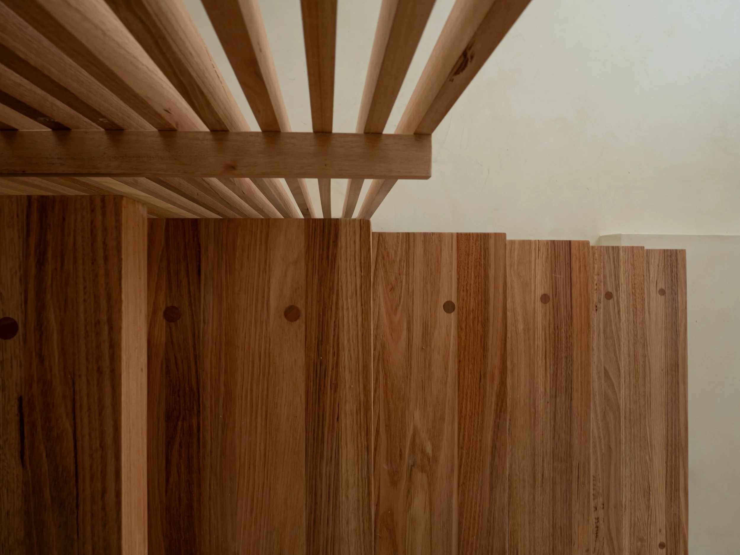 A wooden staircase viewed from above, showing horizontal wooden steps and vertical side panels, with a plain light-colored wall in the background.