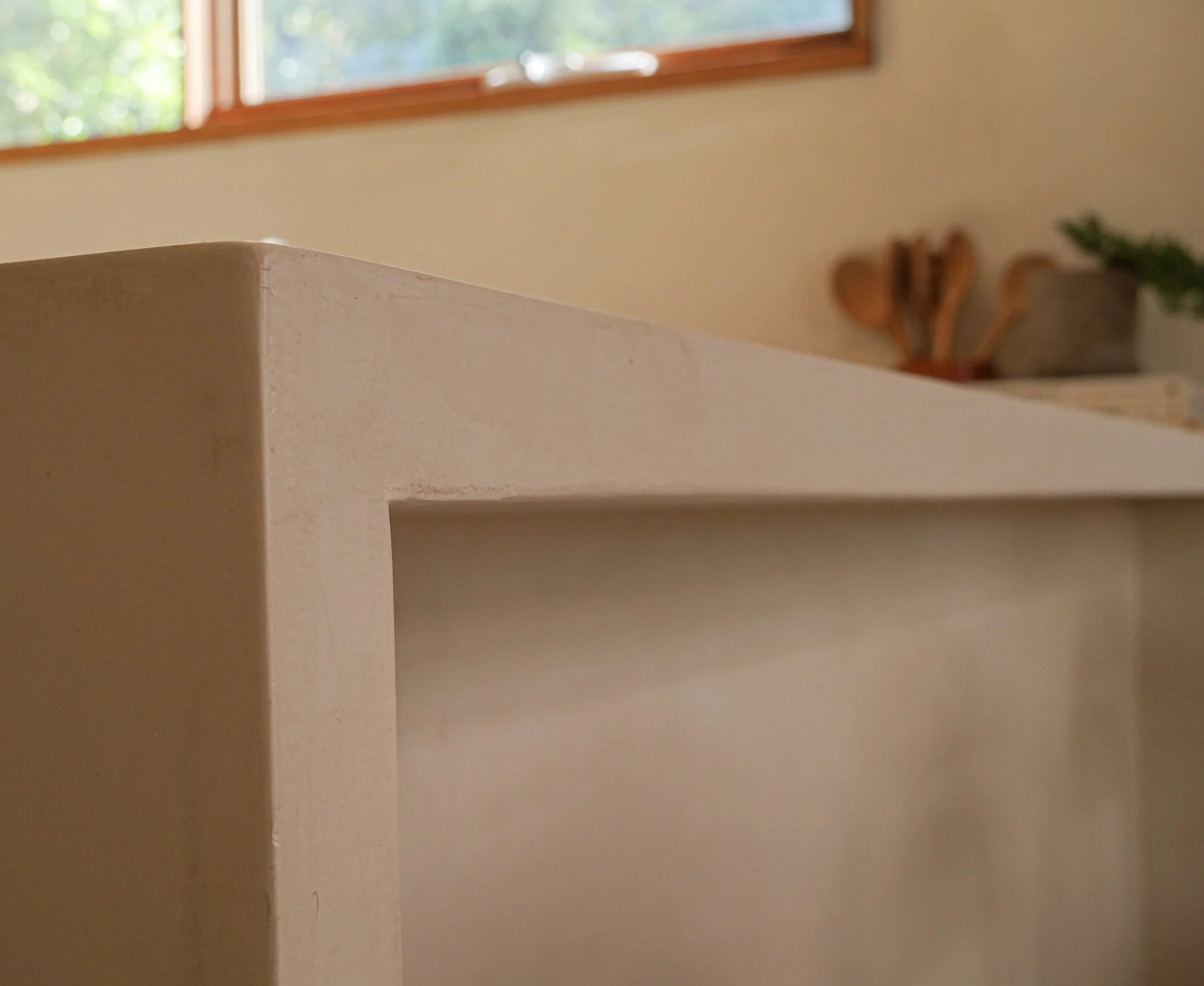 A beige countertop against a wall with a window above it. The window has a wooden frame, and outdoors there are trees visible. There is a small potted plant and a wooden sculpture on the countertop, but the image is blurry.