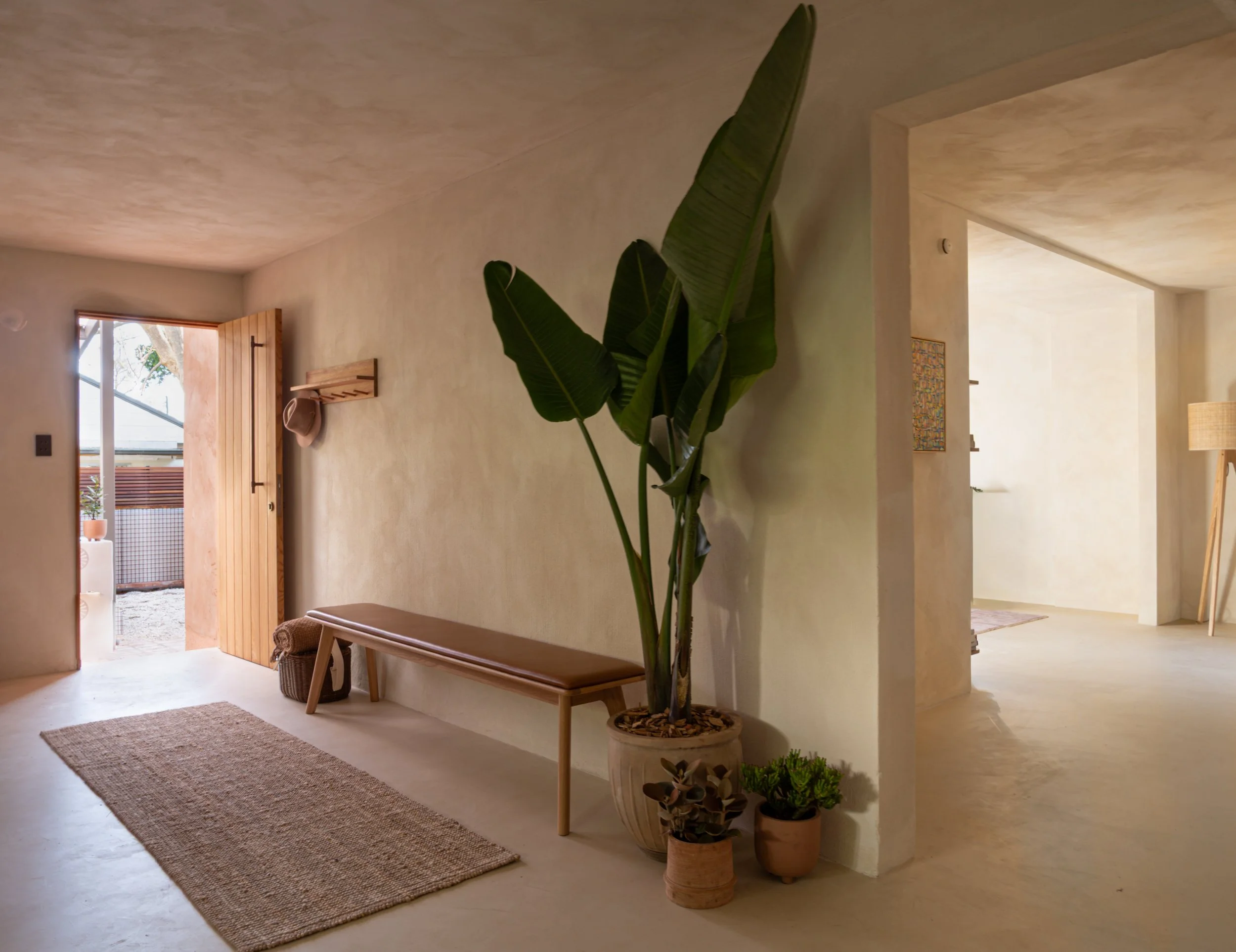 Living room entrance with open wooden door, potted plants, a wooden bench, beige walls, a small woven rug on the floor, and a glimpse of outdoor patio with trees and a lattice fence.