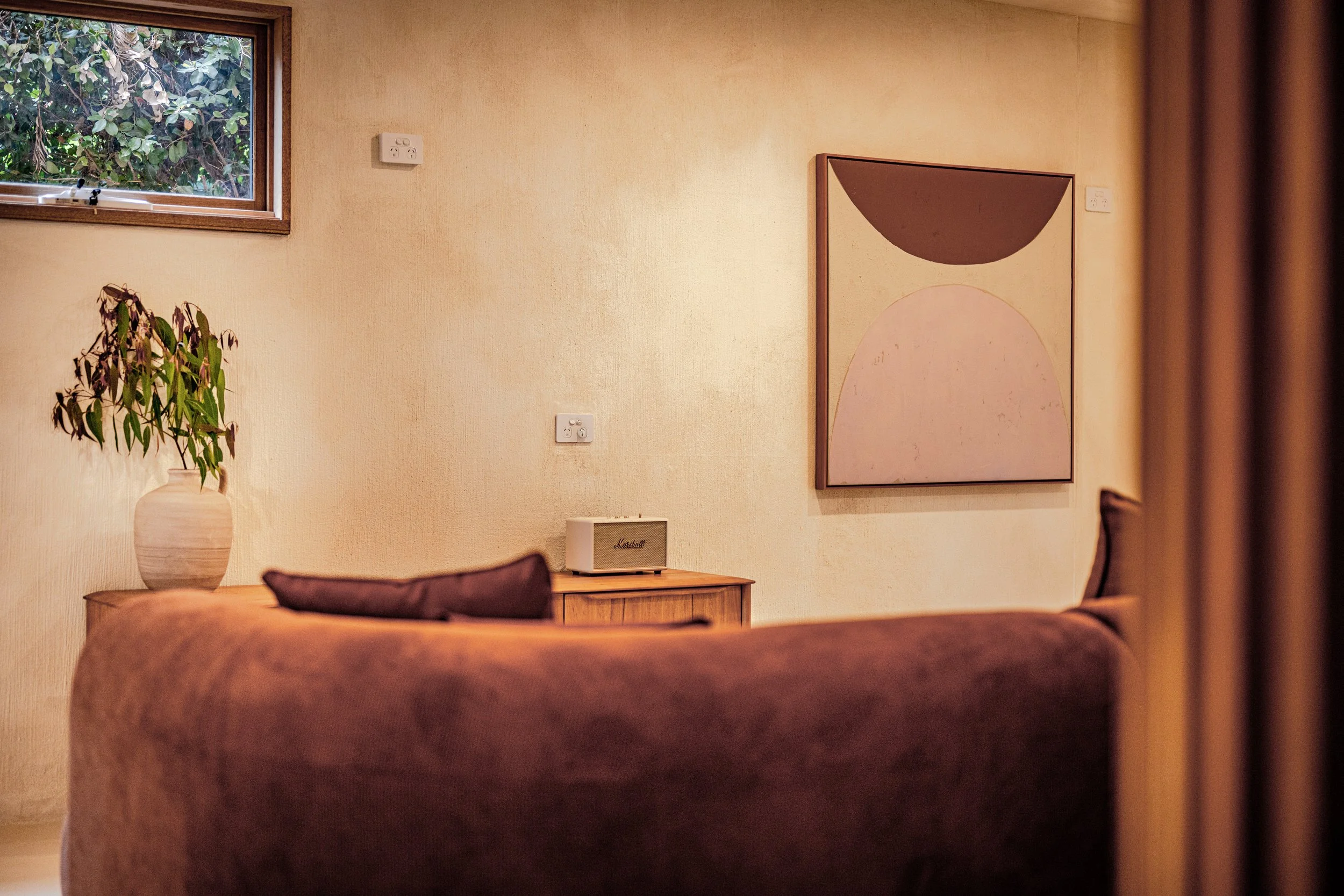 Living room with a window showing outdoor greenery, a potted plant on a wooden table, a framed abstract art piece on the beige wall, and a small speaker on a cabinet.