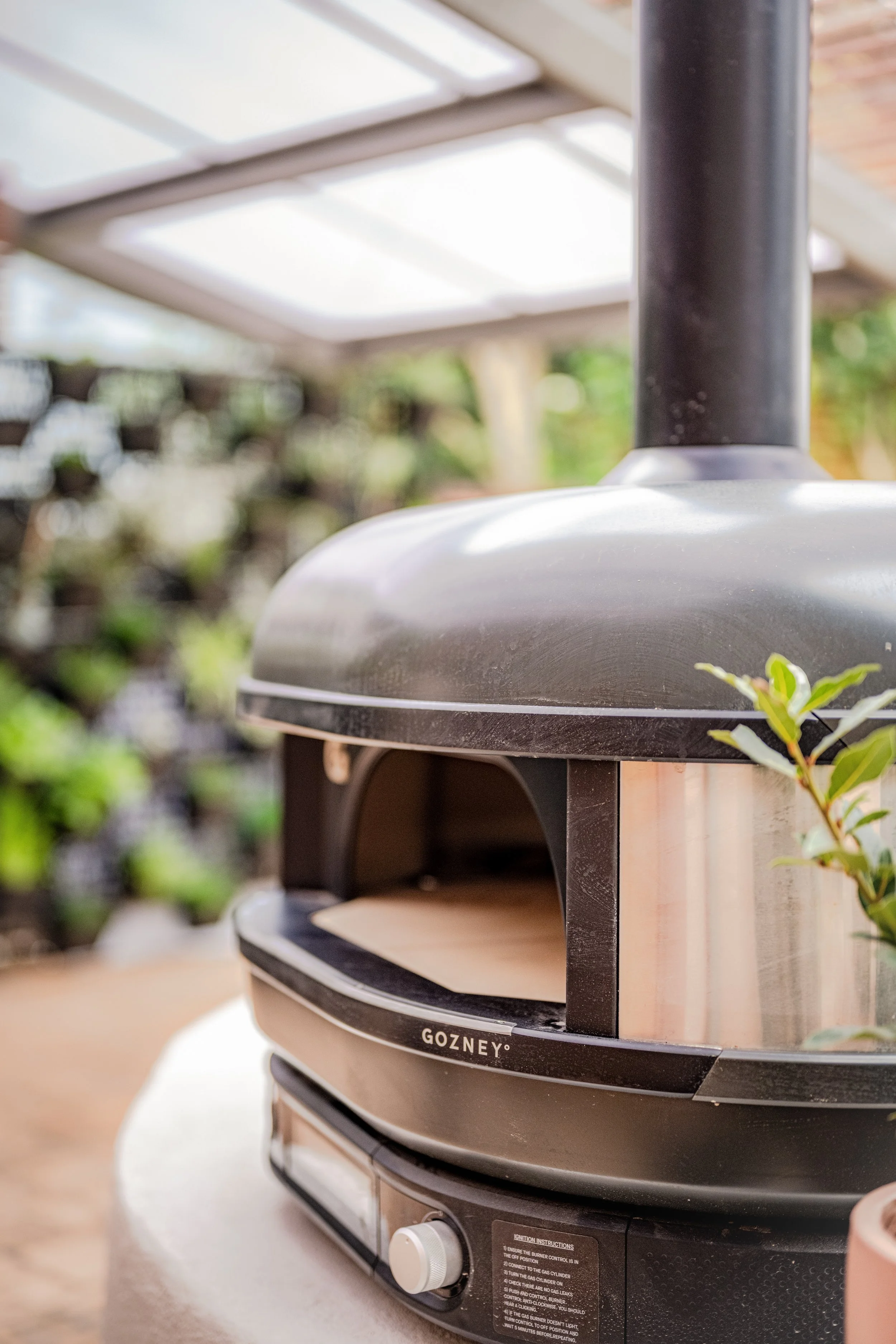 Close-up of a Gozney pizza oven with a wooden exterior, black chimney, and control knob, situated outdoors with plants and a wooden fence in the background.