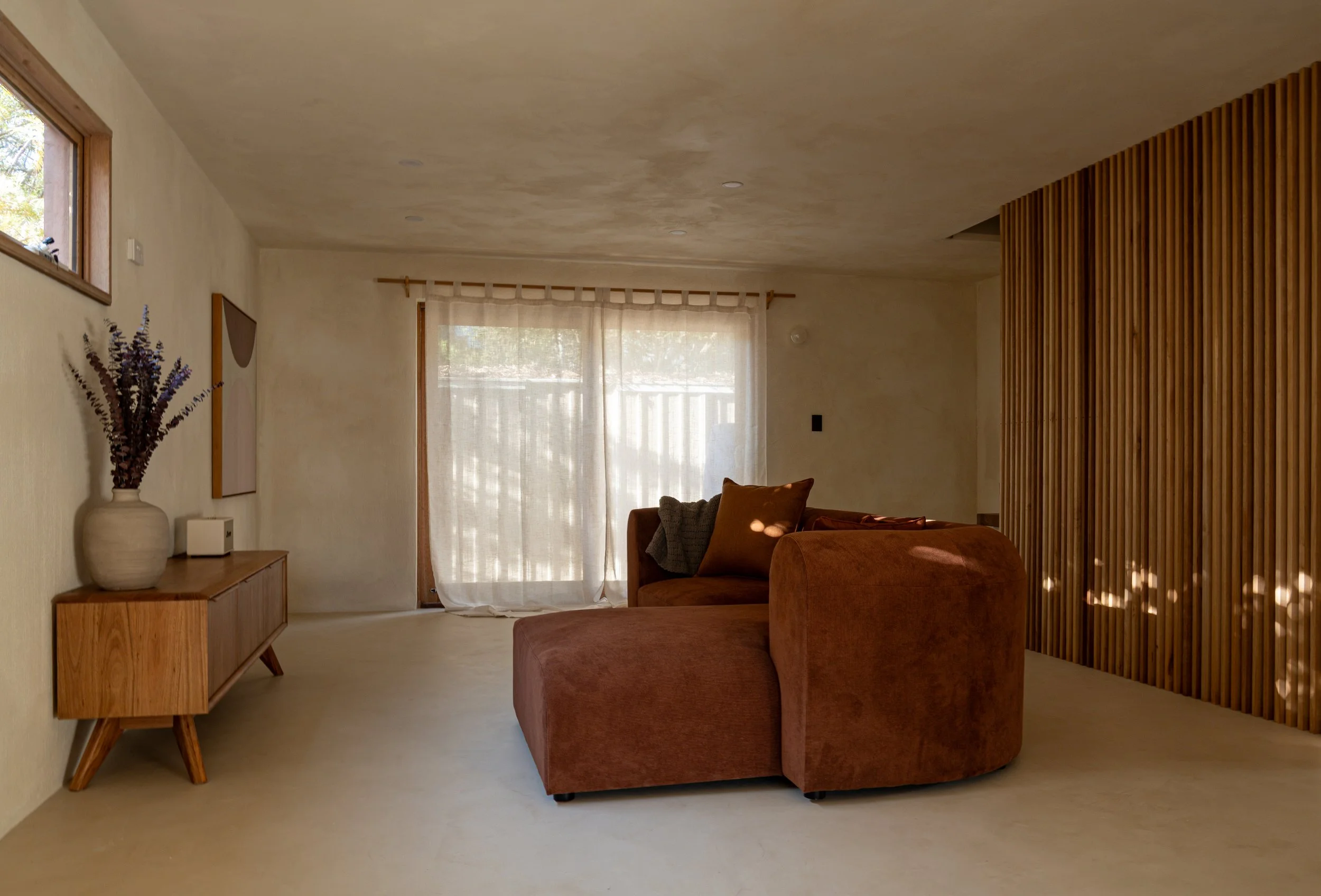 A cozy living room with beige walls and ceiling, a large window with sheer curtains, a wooden sideboard with decorative vase and flowers, a mid-century modern armchair, a sectional sofa with cushions, and a wooden slat room divider.