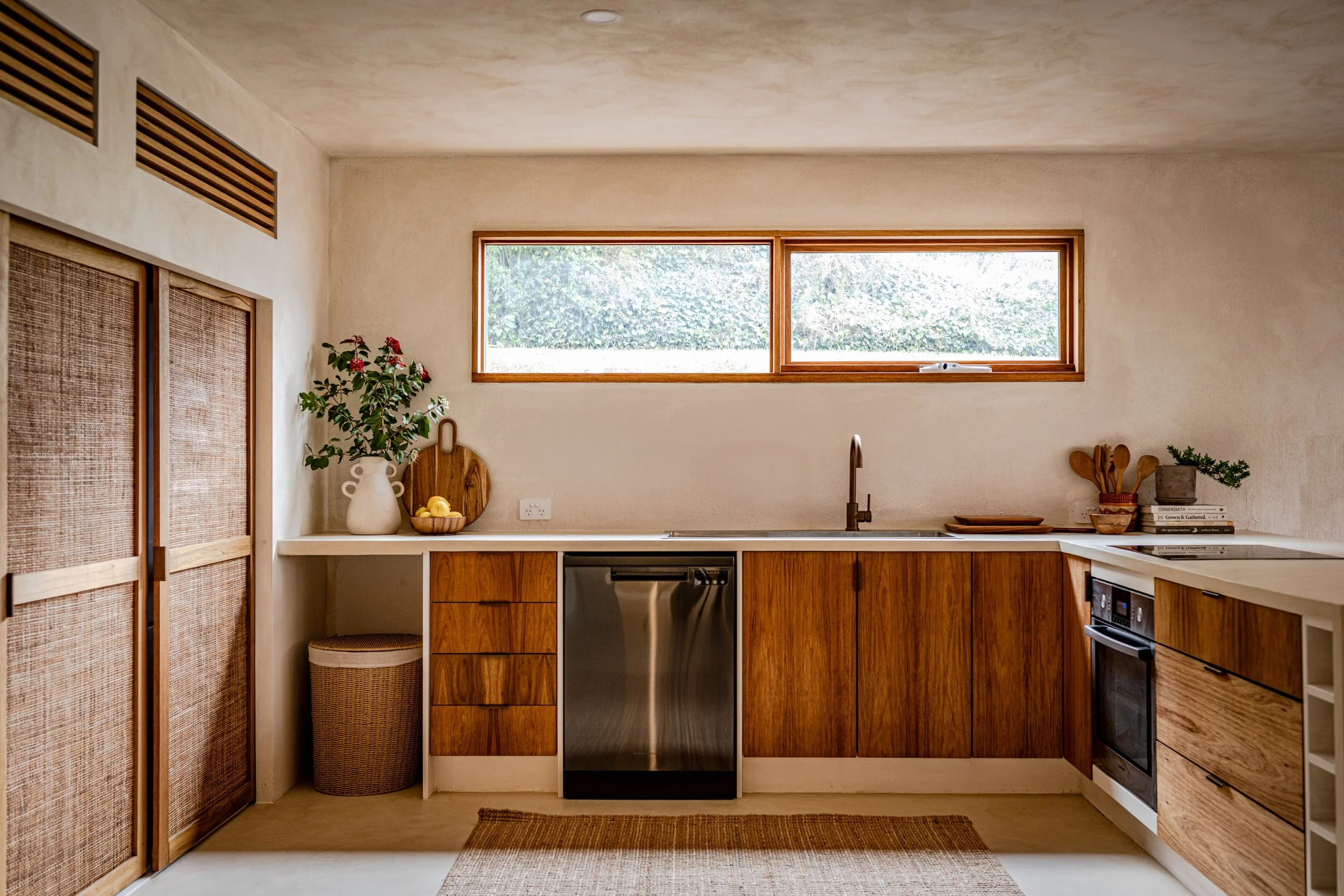 A modern kitchen with wooden cabinets, a large window with a view of green foliage, and various decorative items including a vase with flowers, a basket of lemons, and stacked books.