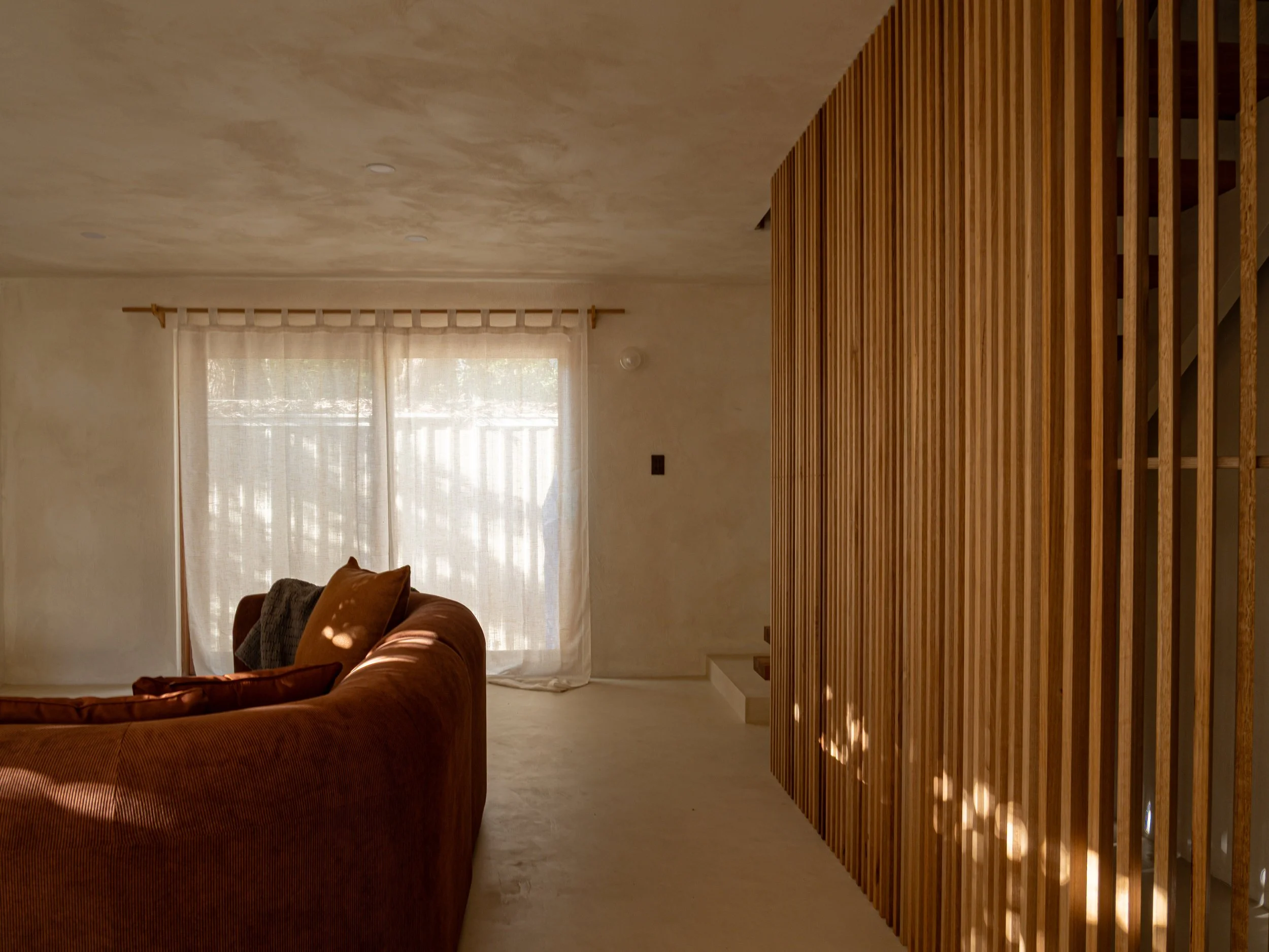 Living room with beige walls, large window with sheer curtains, brown sofa with pillows, and a wooden slat room divider.