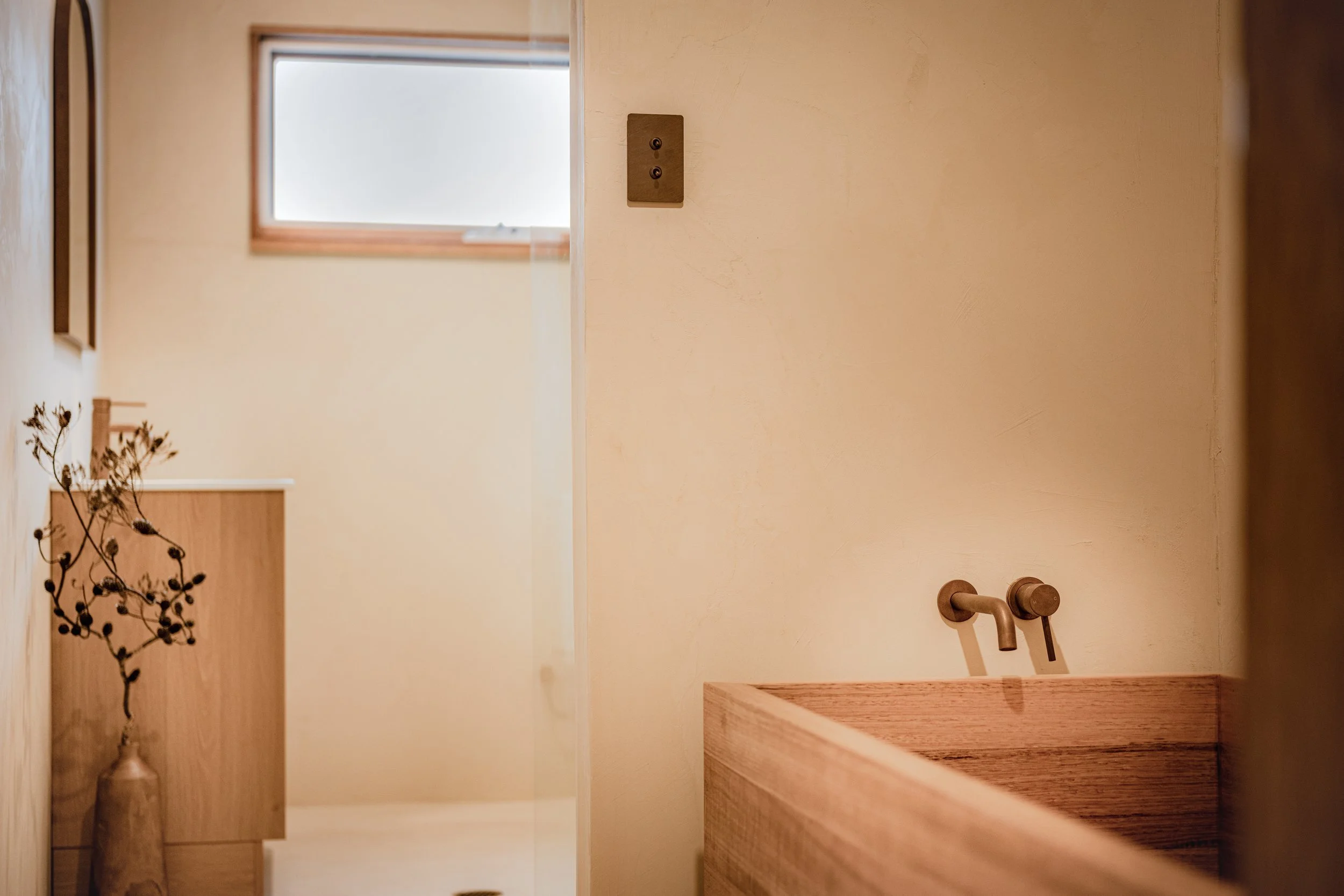 A minimalist bathroom with a beige wall, a small frosted window, a wall-mounted bronze faucet, a wooden countertop, and a dried plant in a large ceramic vase.