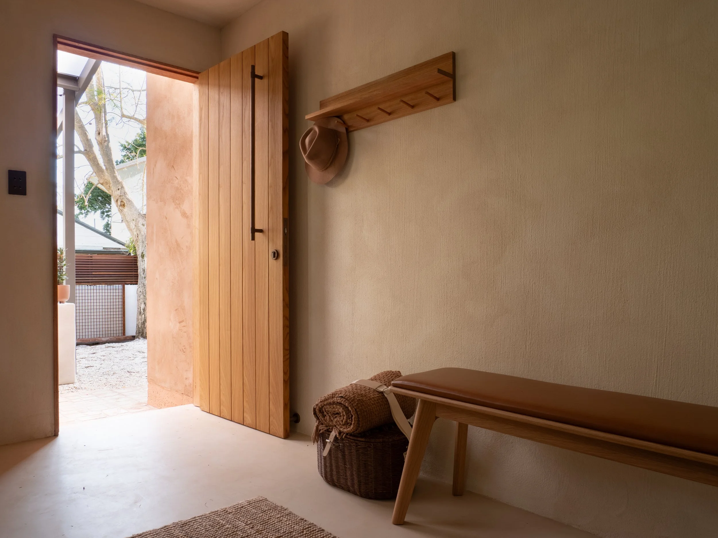 Interior view of a minimalist entryway with a wooden door, a small shelf, a hat hanging, rolled towels, a wicker basket, and a window showing trees outside.