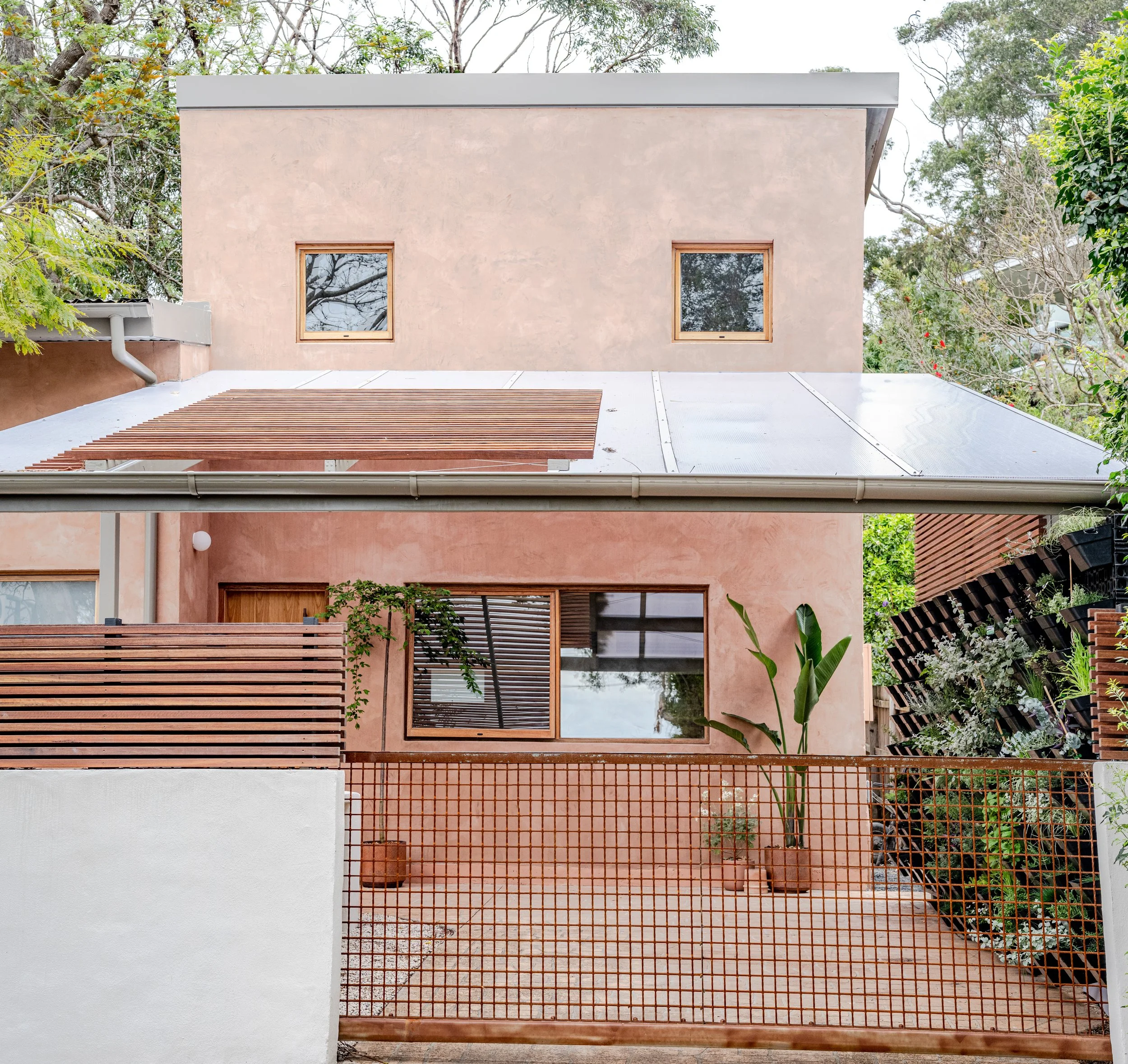 The image shows the upper part of a modern house with two small windows, a flat roof, and surrounded by trees. The house features a mix of pastel pink ochre-colored walls, a white section, and a brown wooden fence with horizontal slats in the foreground. There's also a metal grid gate at the bottom.