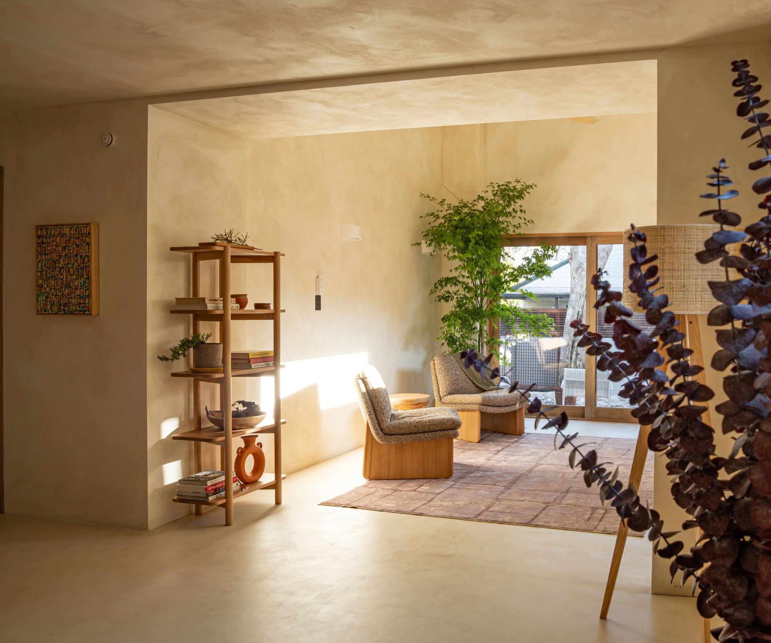 A cozy living room area with beige walls and natural light from large windows. There are two armchairs, a wooden bookshelf with books and decorative items, and large green plants near the windows. The floor has a light-colored carpet and brick-patter