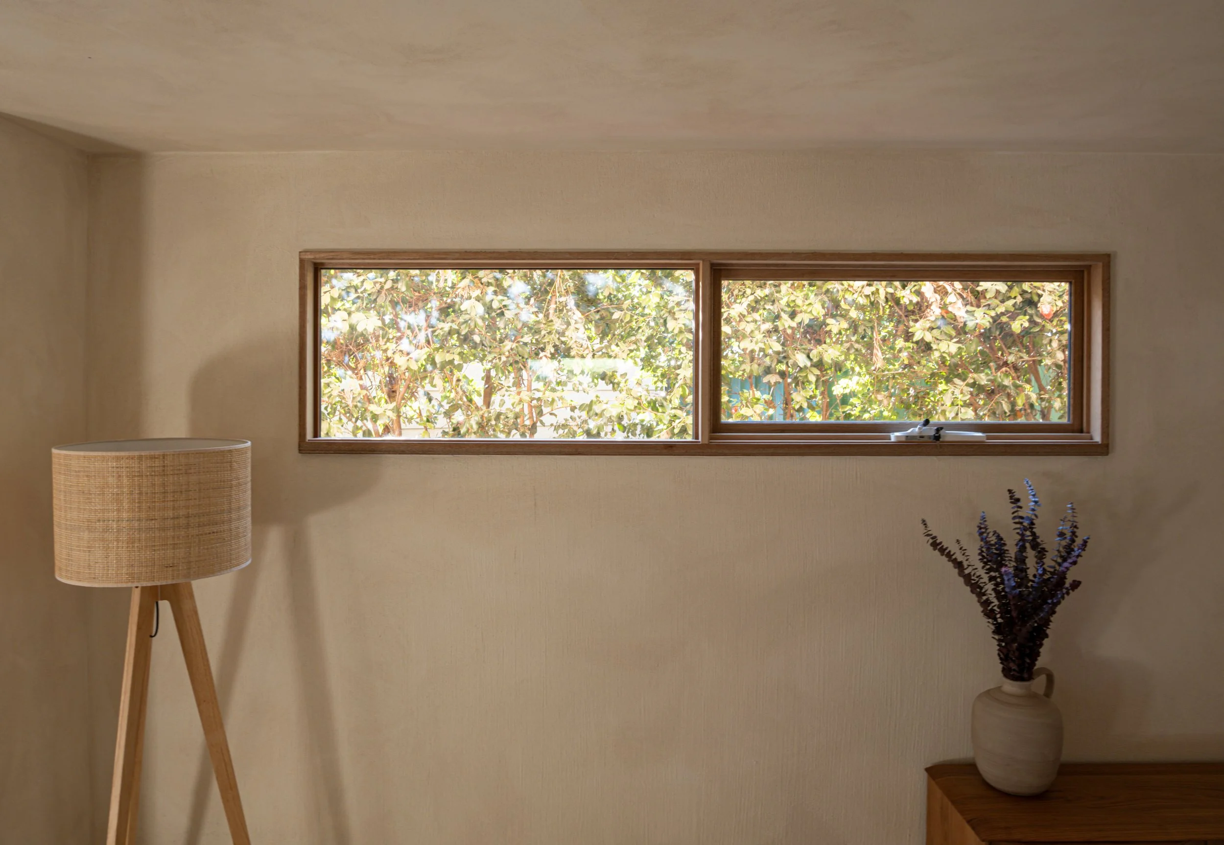 Interior of a room with a horizontal window showing trees outside, a beige lampshade on a wooden tripod, a wooden side table with a white vase containing purple flowers, and beige walls and ceiling.