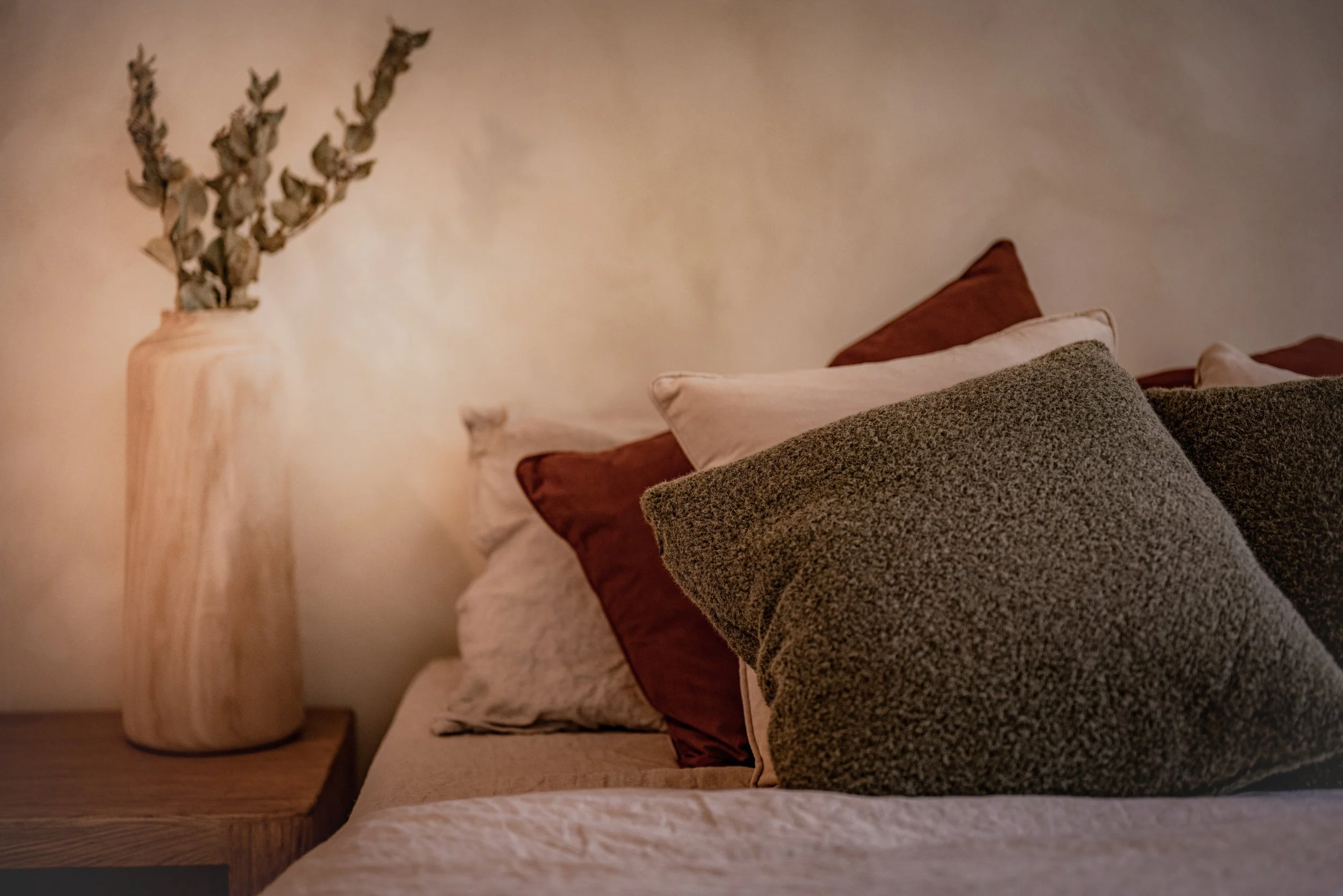 Close-up of a bedroom with a beige bed and multiple pillows, a wooden nightstand, and a tall, light-colored wooden lamp with a dried plant arrangement